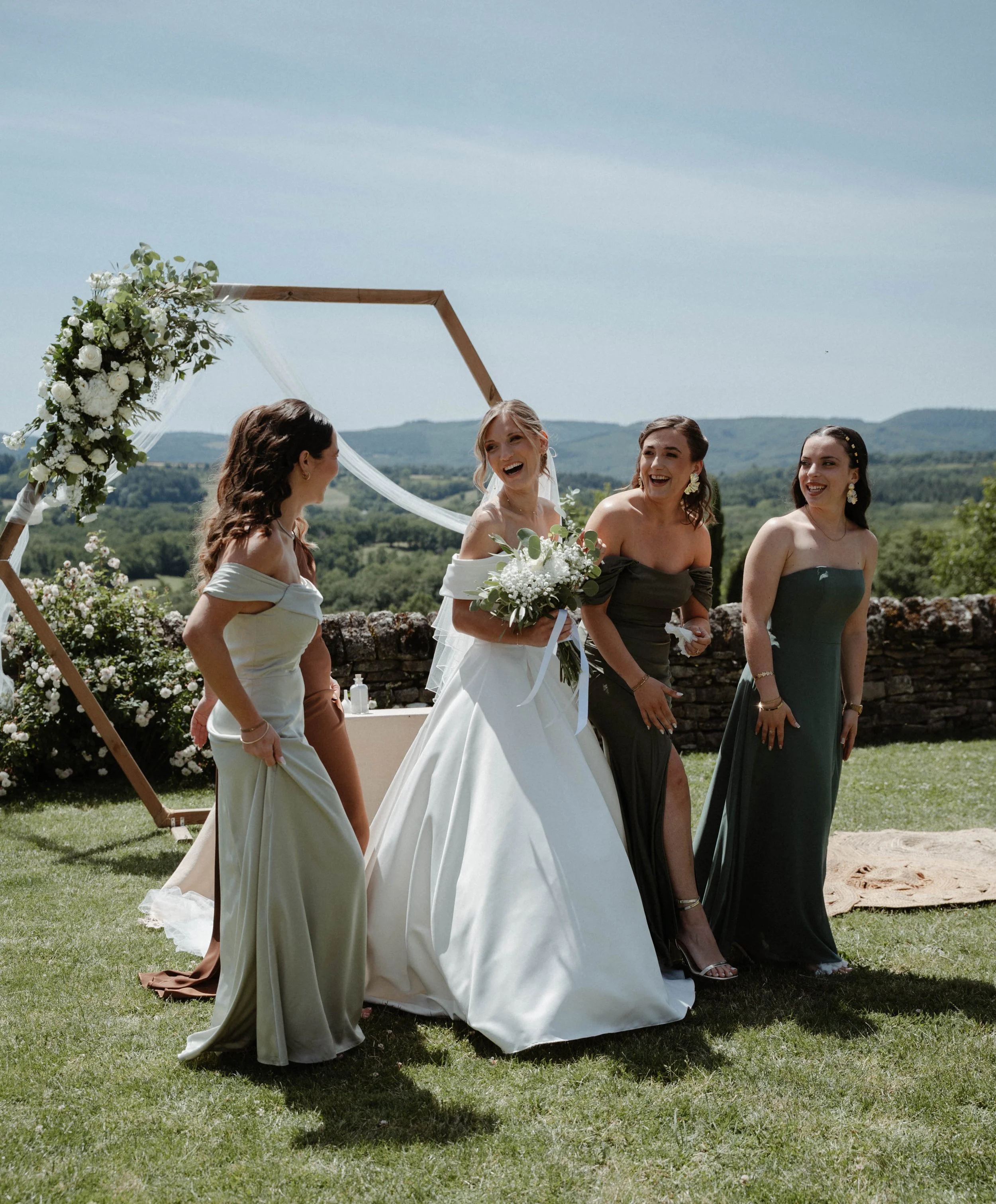 Quatre femmes en robes de mariage et de soirée, lors d'un mariage en plein air, rire et discuter sous un ciel bleu avec un paysage de collines en arrière-plan.