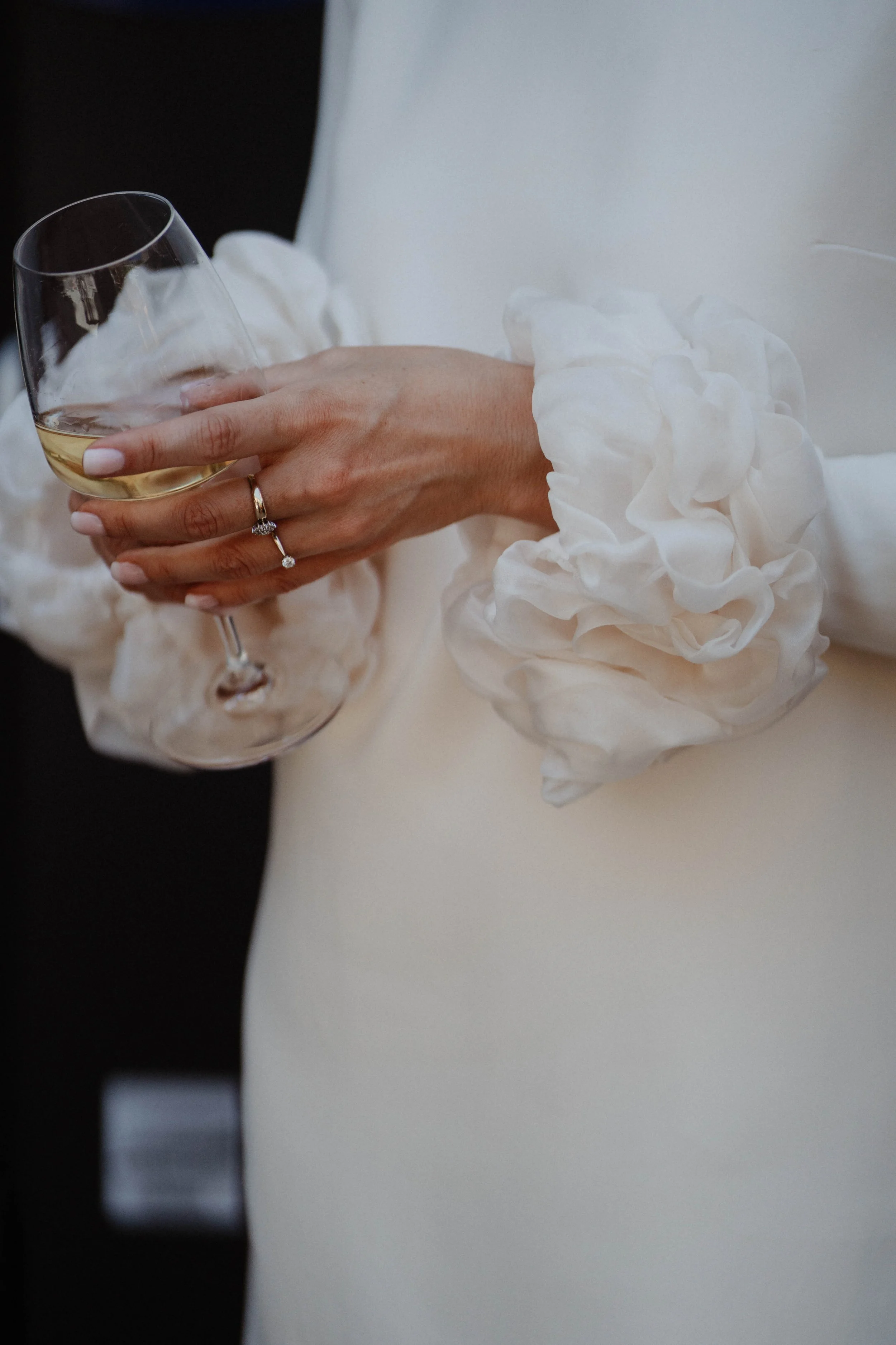 Une main portant une bague tient un verre de vin blanc. La main est vêtue d'une robe avec des manches en tissu froissé blanc.