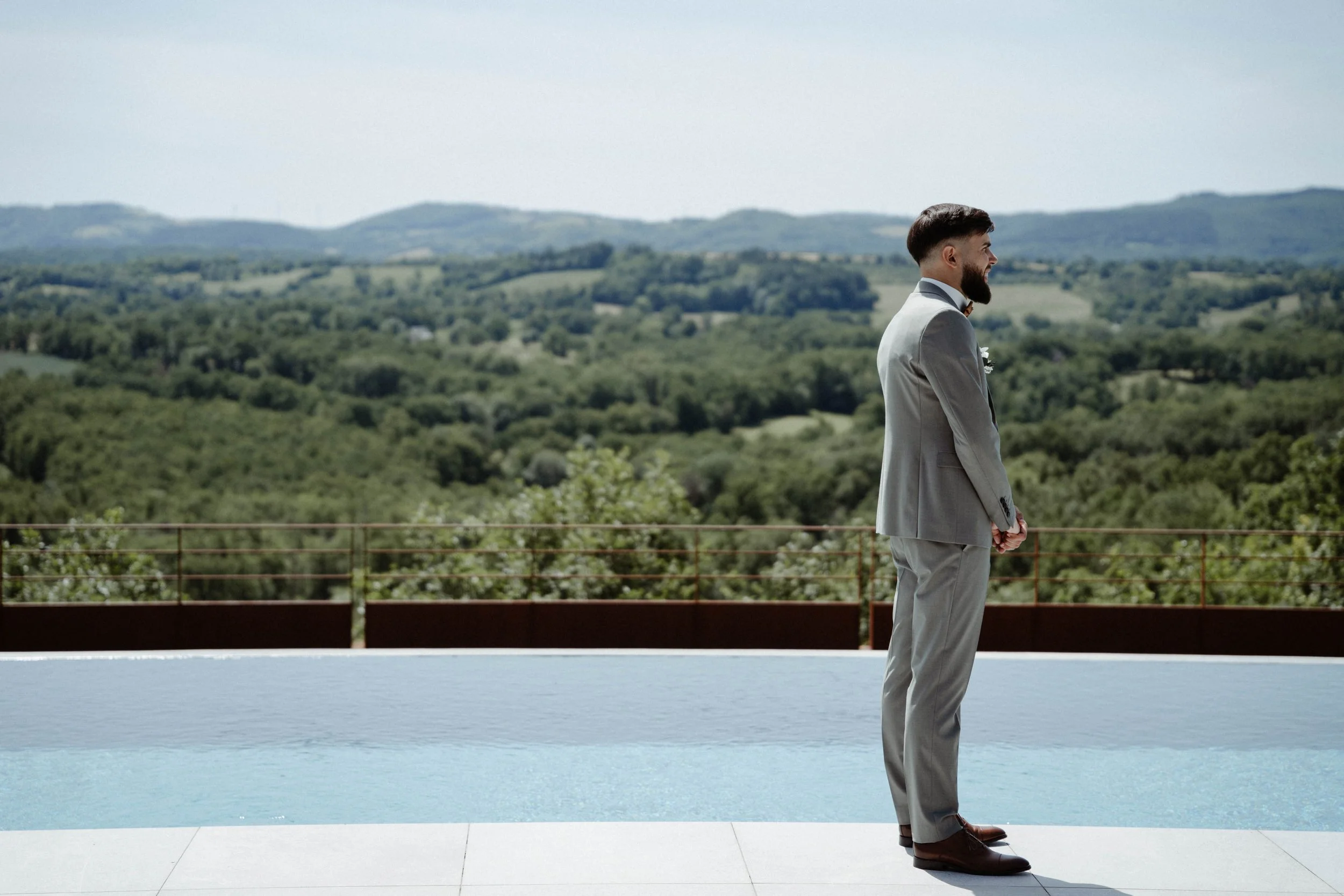 Un homme en costume gris, barbe, porte une coiffure moderne, se tient debout sur une terrasse avec vue sur un paysage de collines verdoyantes et un ciel clair.