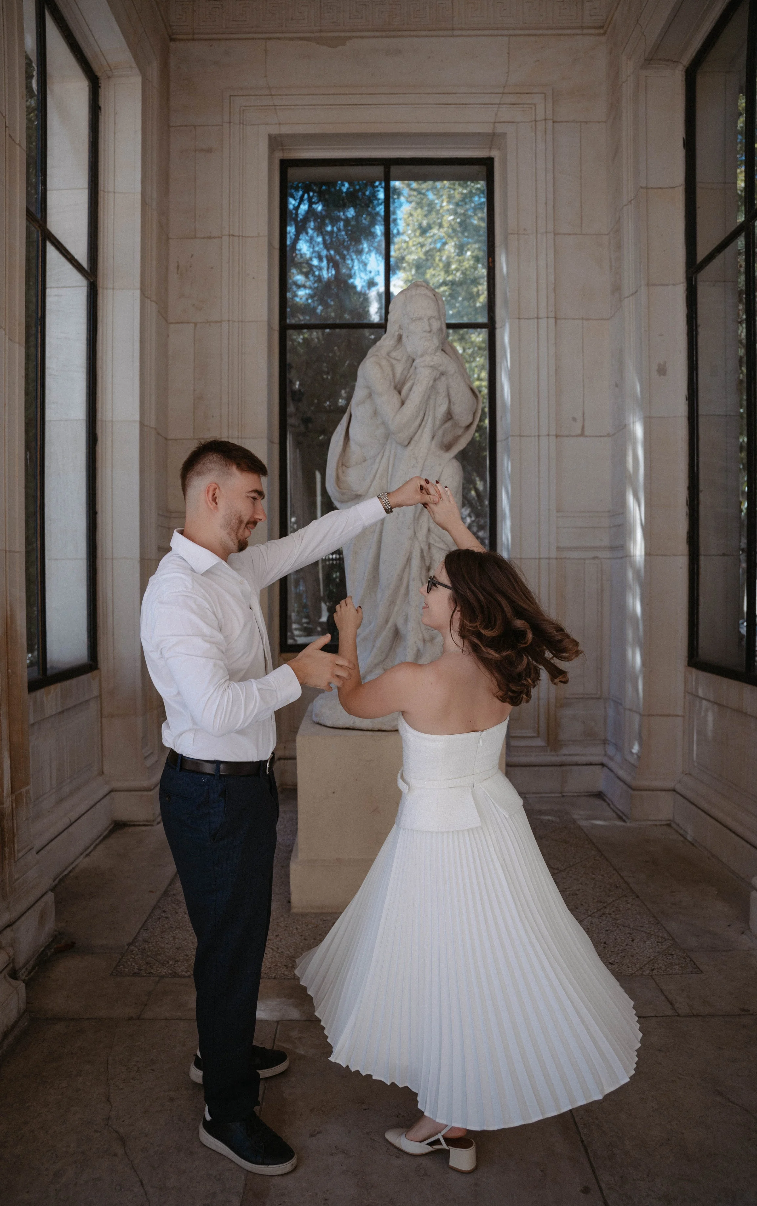Un couple danse dans une salle ornée de sculptures en marbre, avec une grande fenêtre laissant entrer la lumière naturelle.