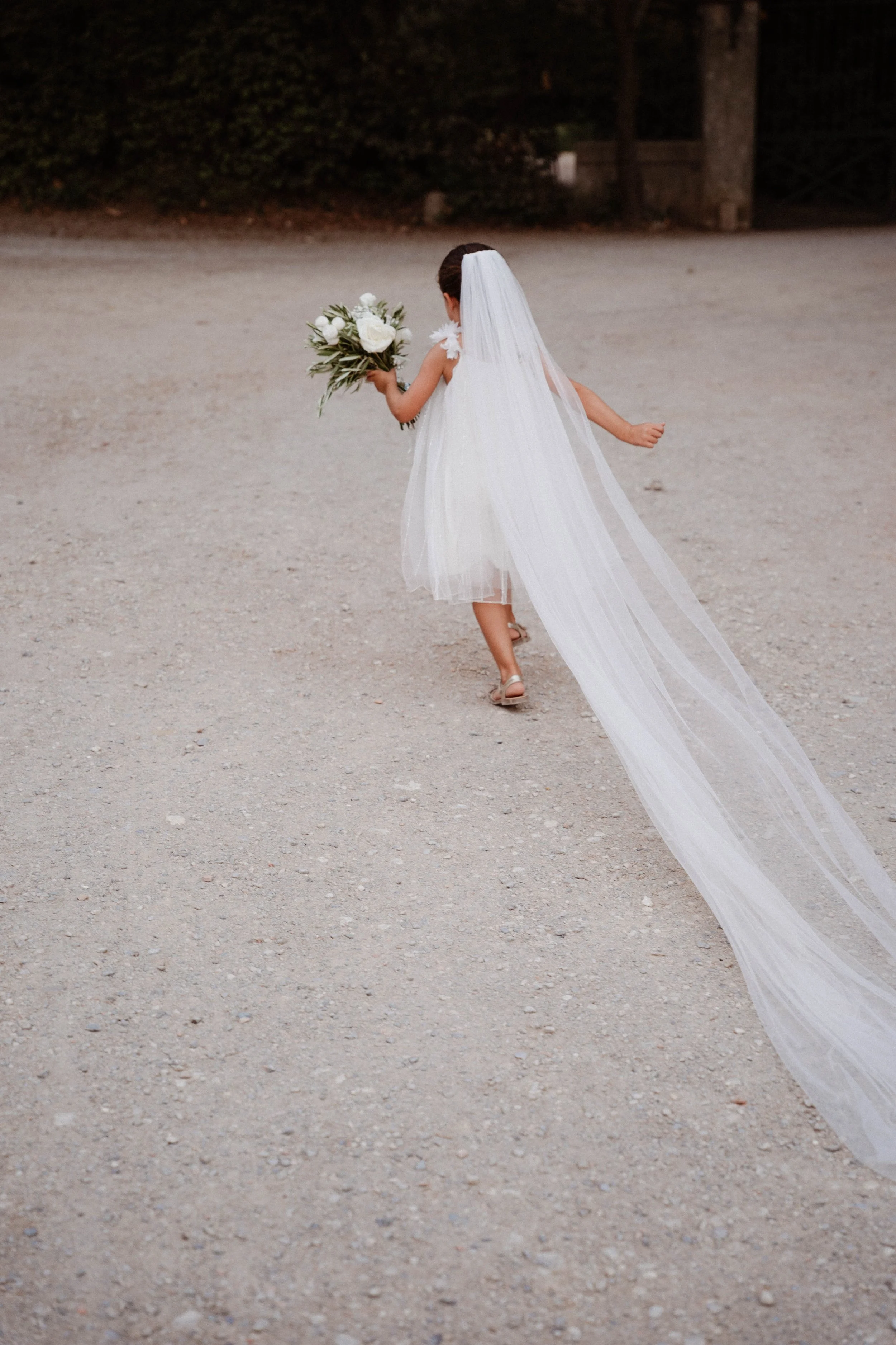 Une jeune femme en robe de mariée avec un voile, tenant un bouquet de fleurs blanches, marche sur un chemin en extérieur.