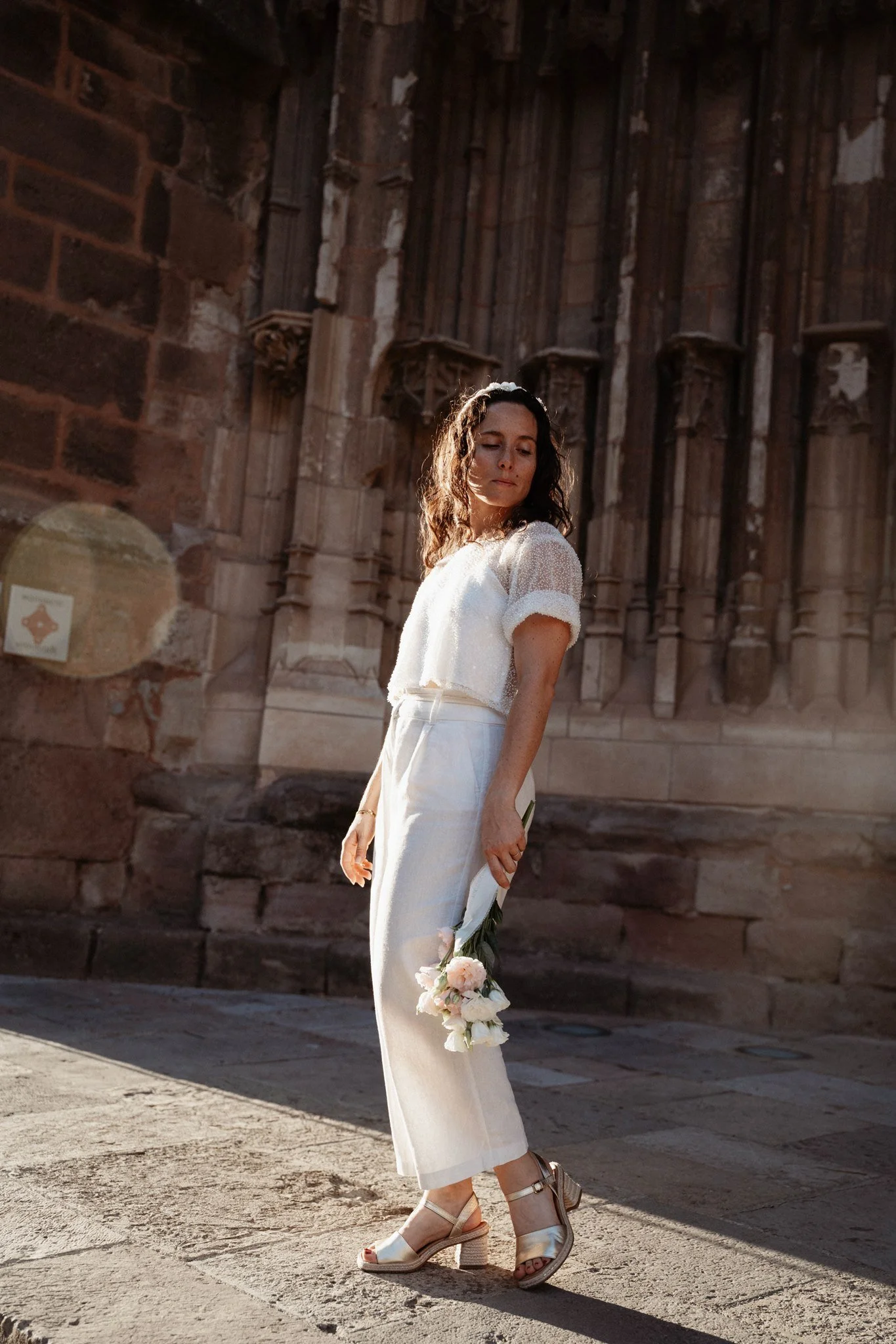 Une femme habillée en blanc avec des hauteurs, portant des chaussures à talons, tient un bouquet de fleurs, se tenant devant une ancienne structure en pierre, avec une expression calme et une lumière chaleureuse. L'arrière-plan est constitué de murs 