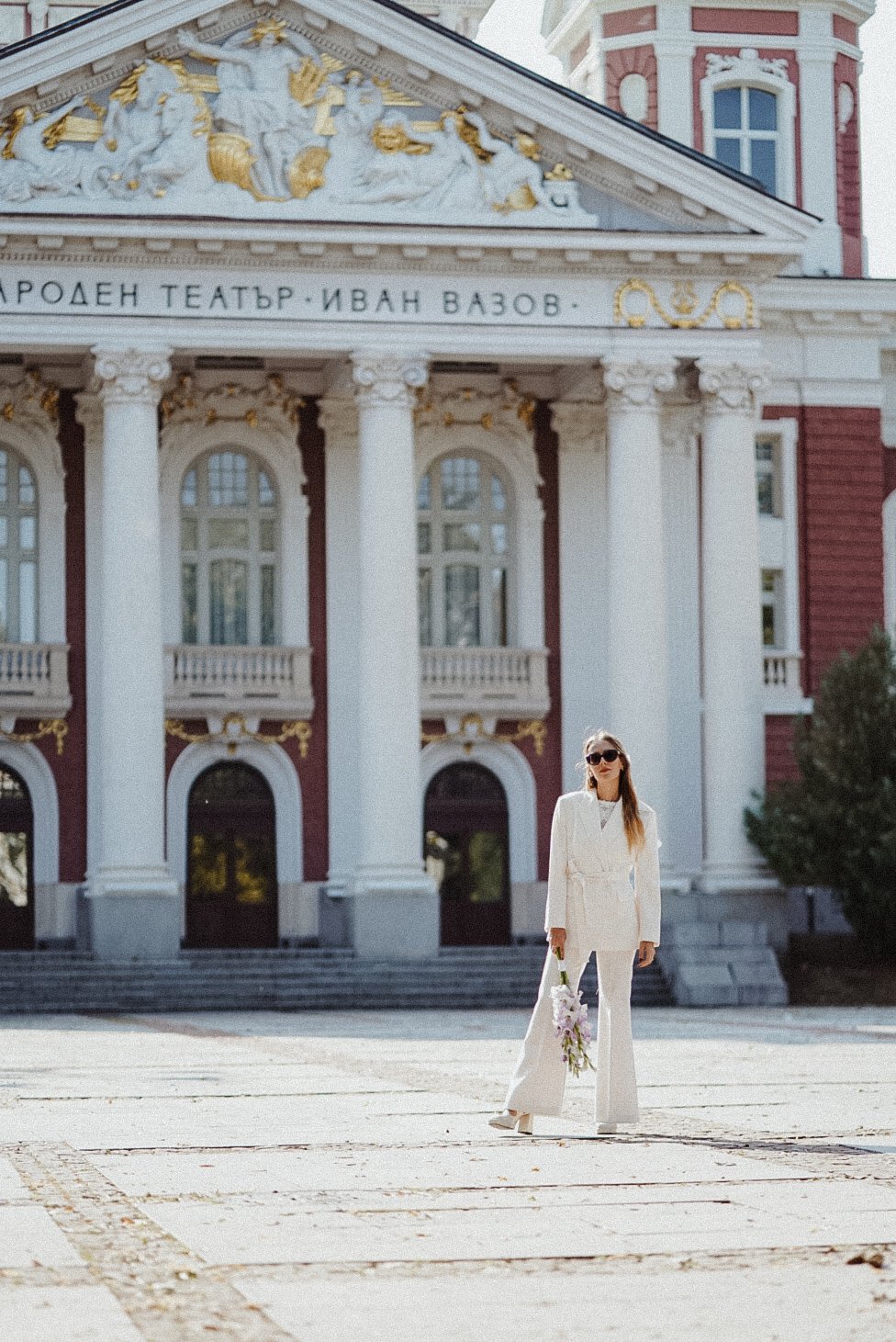 Une femme habillée en blanc avec des lunettes de soleil, tenant un bouquet de fleurs, se tenant devant un bâtiment historique avec des colonnes et une façade ornée.