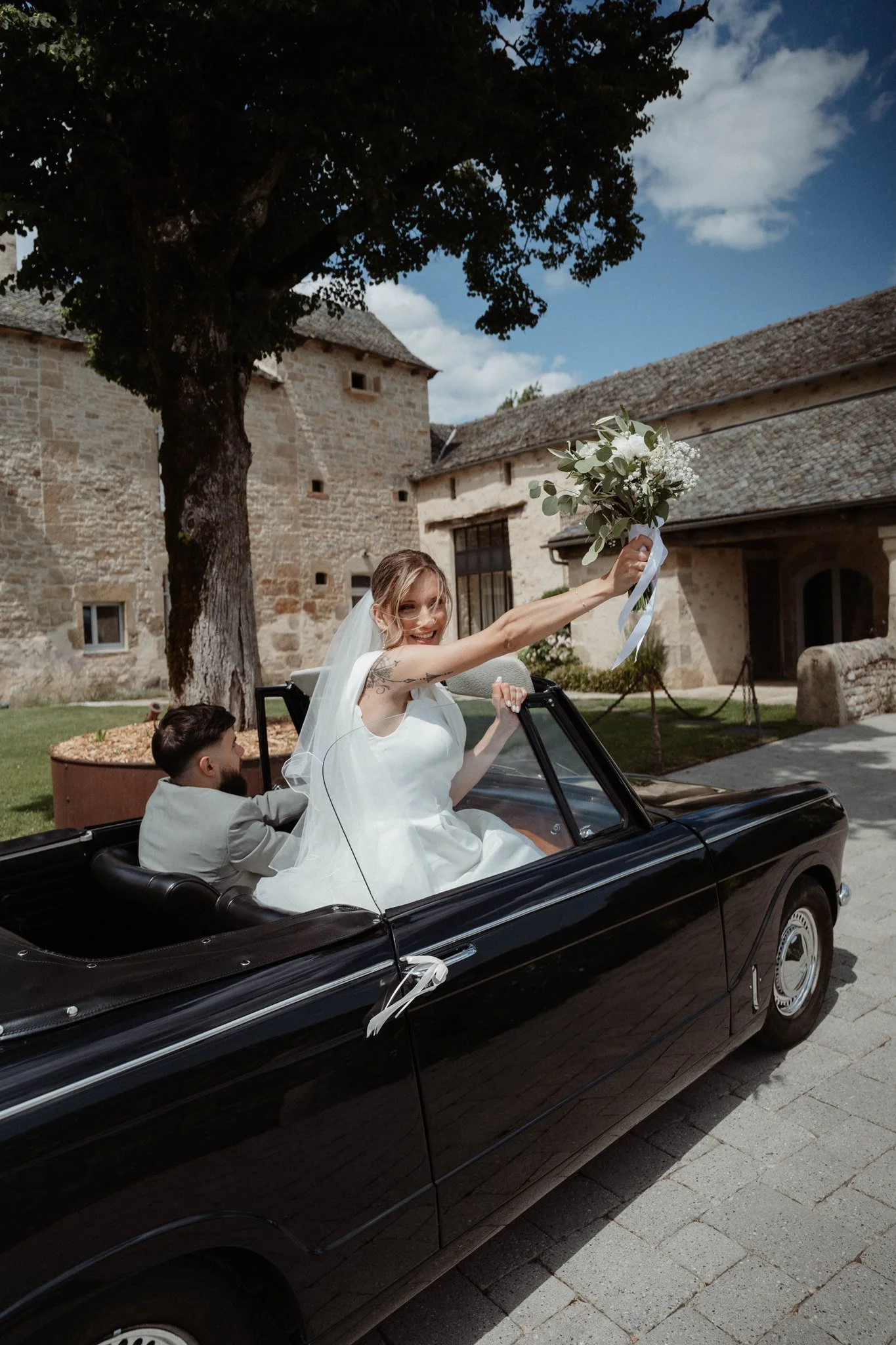 Une mariée joyeuse dans une voiture de mariage noire, tenant un bouquet de fleurs blanches, avec deux personnes dans la voiture, en plein air devant un bâtiment en pierre.