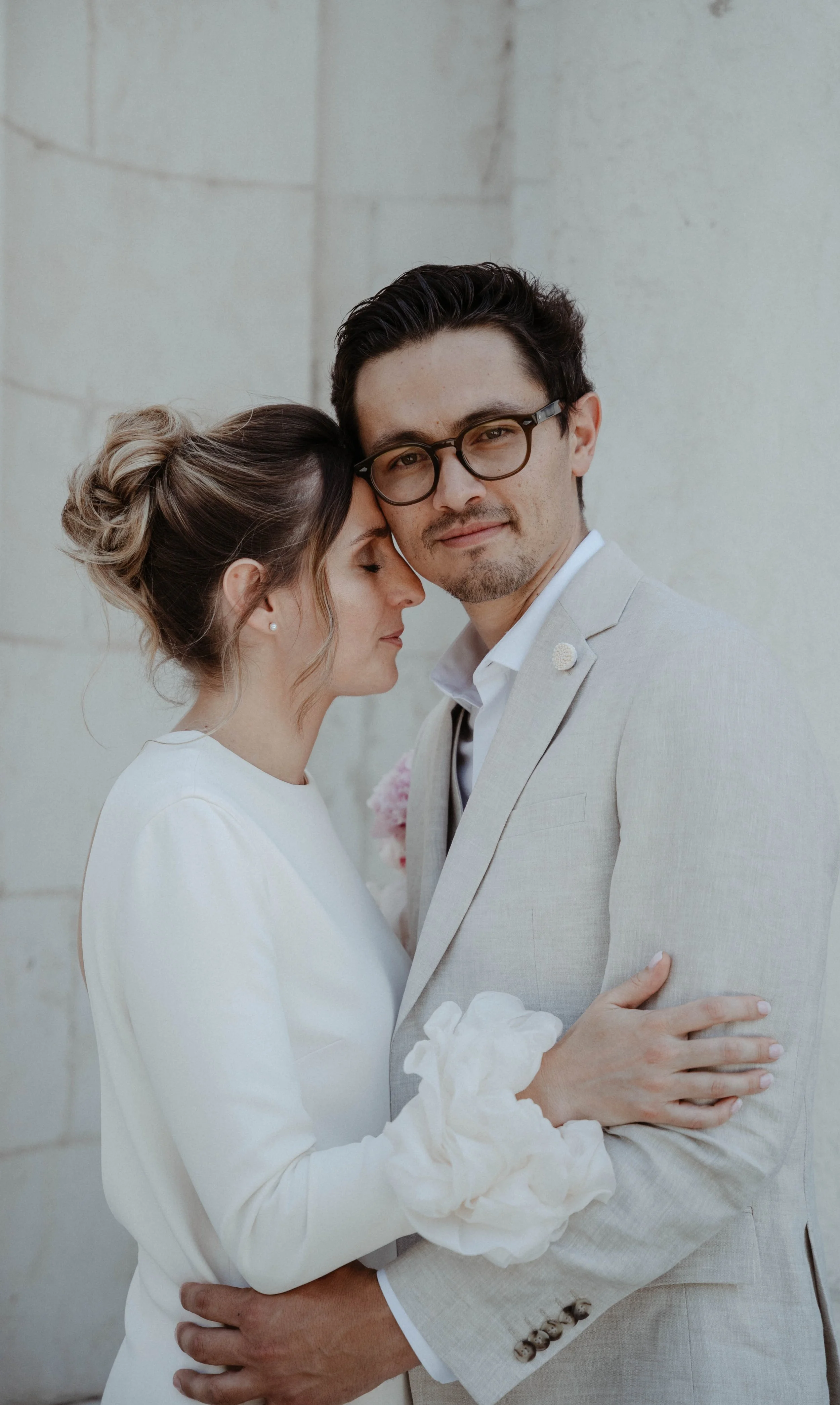 Un couple en tenue de mariage se tient contre un mur en pierre, se regardant tendrement, la femme a les yeux fermés et le homme porte des lunettes et regarde vers l'objectif.