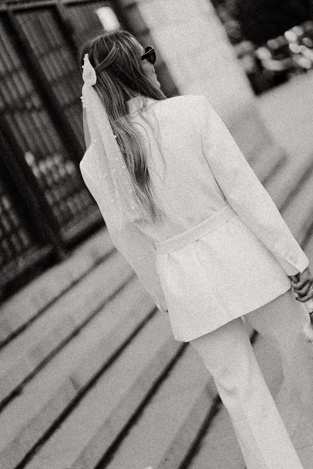 Femme portant un tailleur blanc avec veste et jupe, vue de dos, marchant dans une rue urbaine.