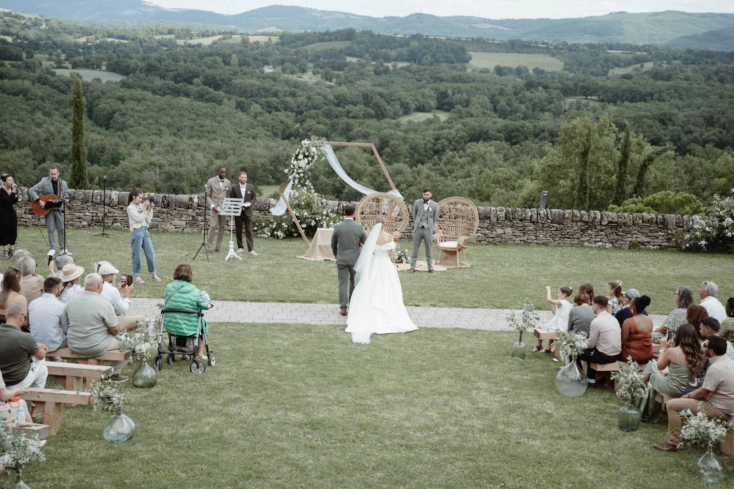 Une cérémonie de mariage en plein air avec des invités assis, un couple marié, un officiant, et un groupe de musiciens, dans un paysage verdoyant avec des collines à l'arrière-plan.
