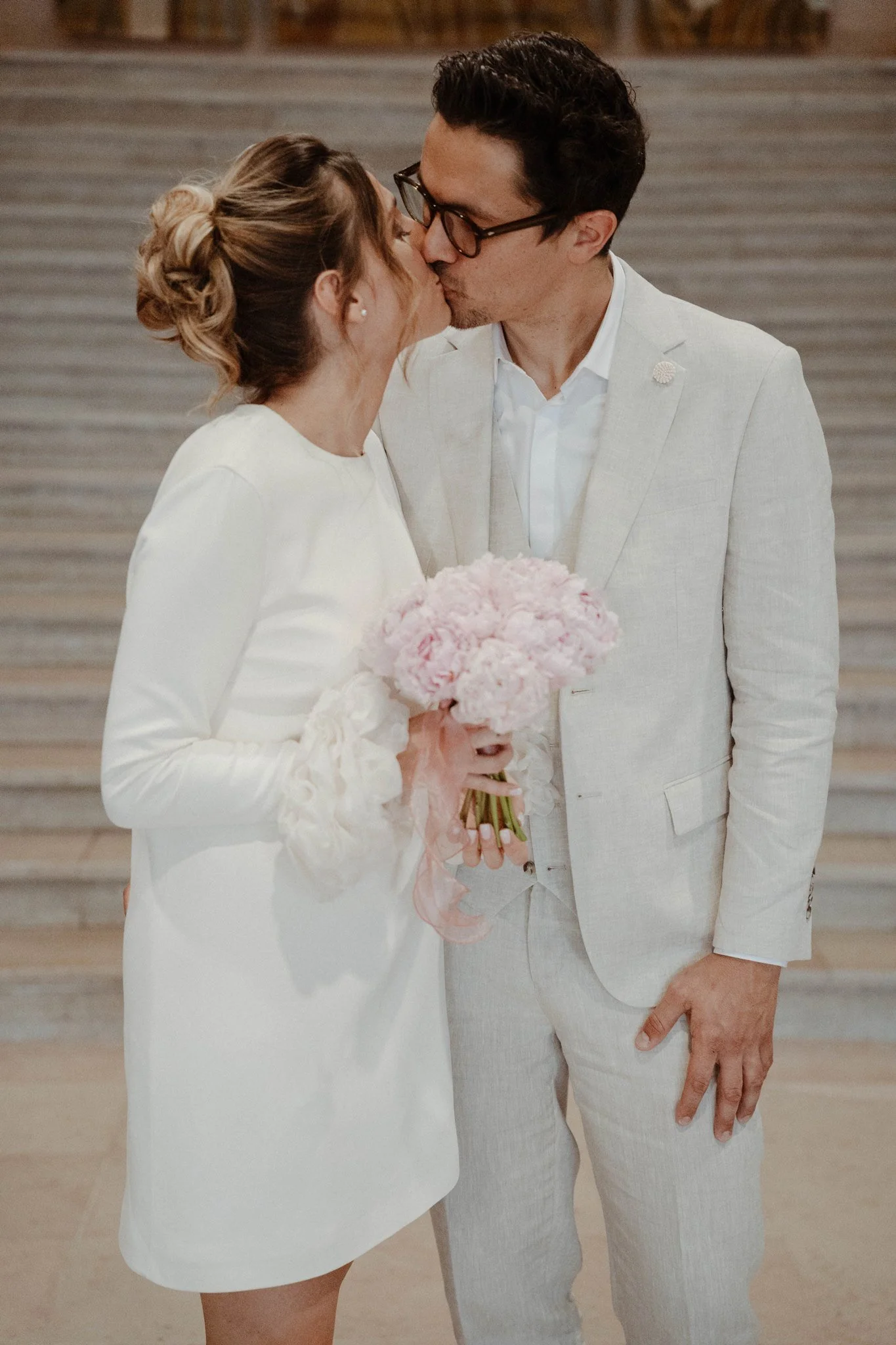 Un couple en tenue de mariage s'embrasse lors de leur mariage civil. La femme tient un bouquet de fleurs roses et blanches, et ils sont en face l'un de l'autre dans un lieu intérieur.