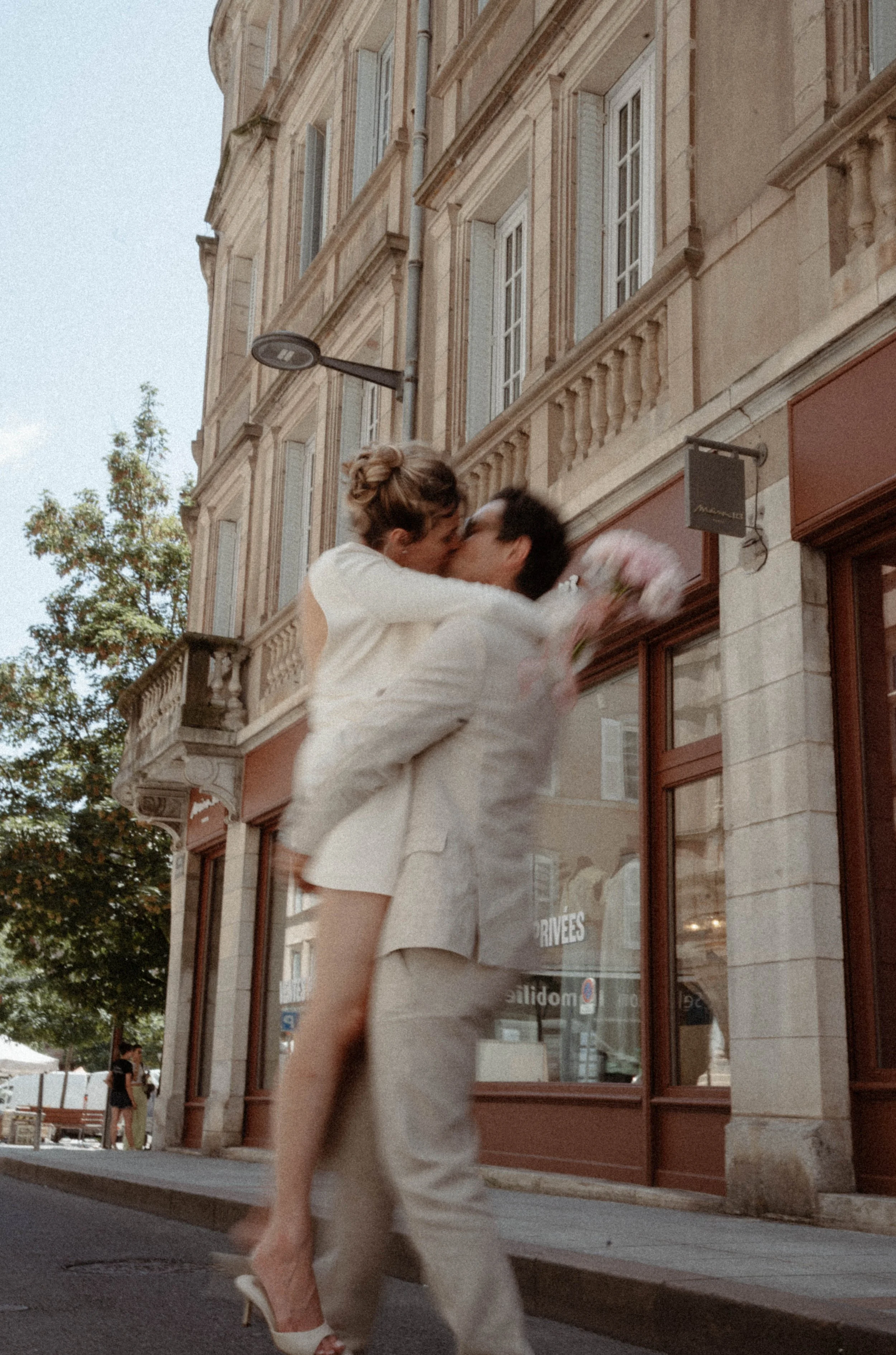 Deux personnes, une femme en robe blanche et un homme en costume clair, s'embrassent devant un bâtiment en pierre ancienne, avec un balcon et des fenêtres à volets bleus.