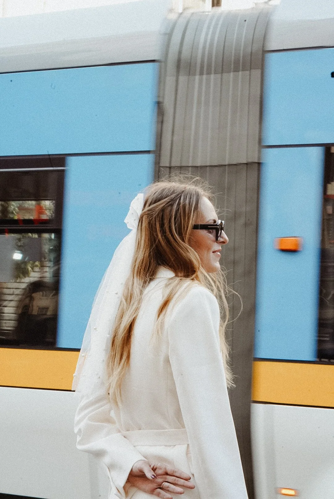 Une femme en costume blanc marche devant un tramway bleu.