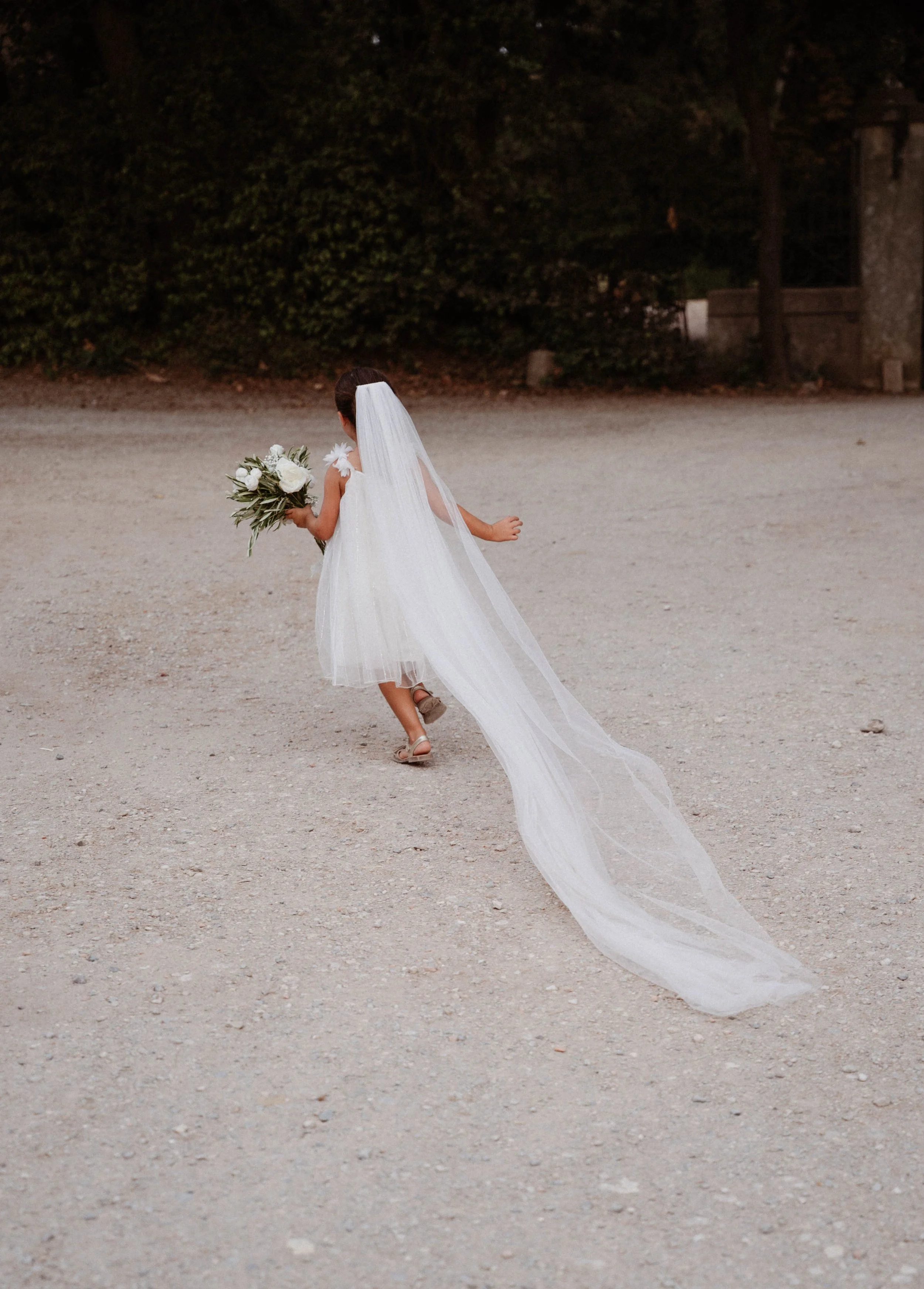 Jeune fille en robe de mariage blanche avec longue voile, tenant un bouquet de fleurs blanches, marchant sur un chemin en terre, vue de dos, avec forêt en arrière-plan.