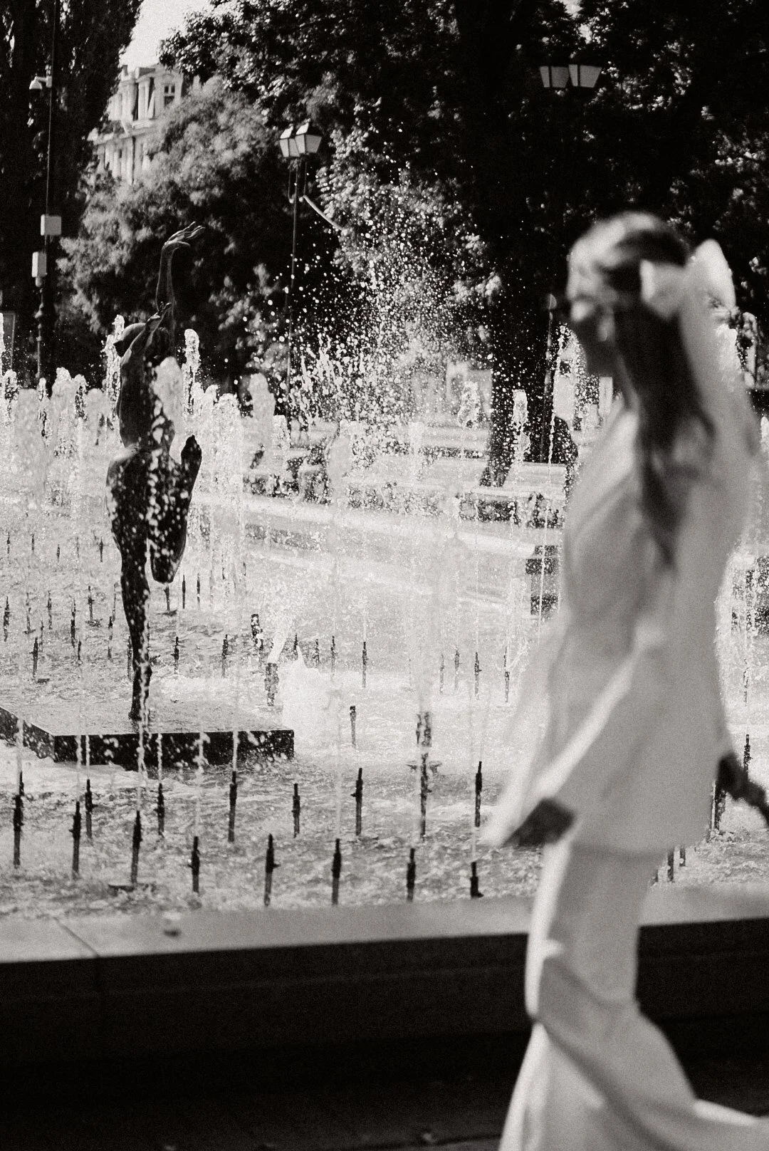 Une fontaine publique avec des statues et des jets d'eau, vue à travers une fenêtre ou une vitre, avec une femme floue en premier plan. L'image est en noir et blanc.
