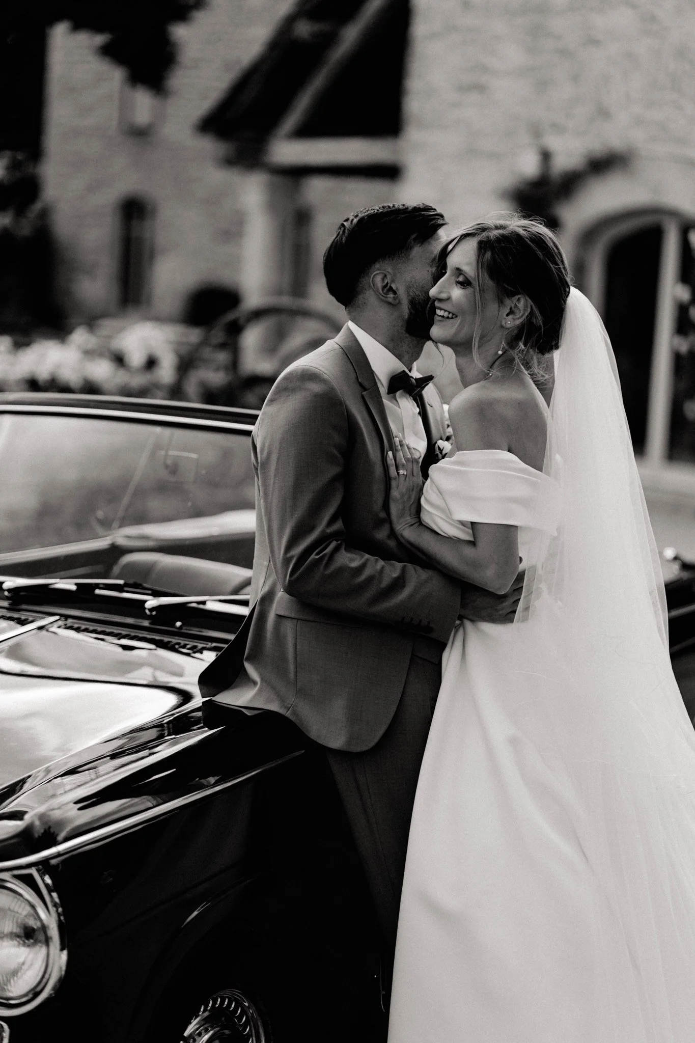 Un couple de mariés souriant, debout près d'une voiture noire classique, avec un bâtiment en arrière-plan, lors d'un mariage.