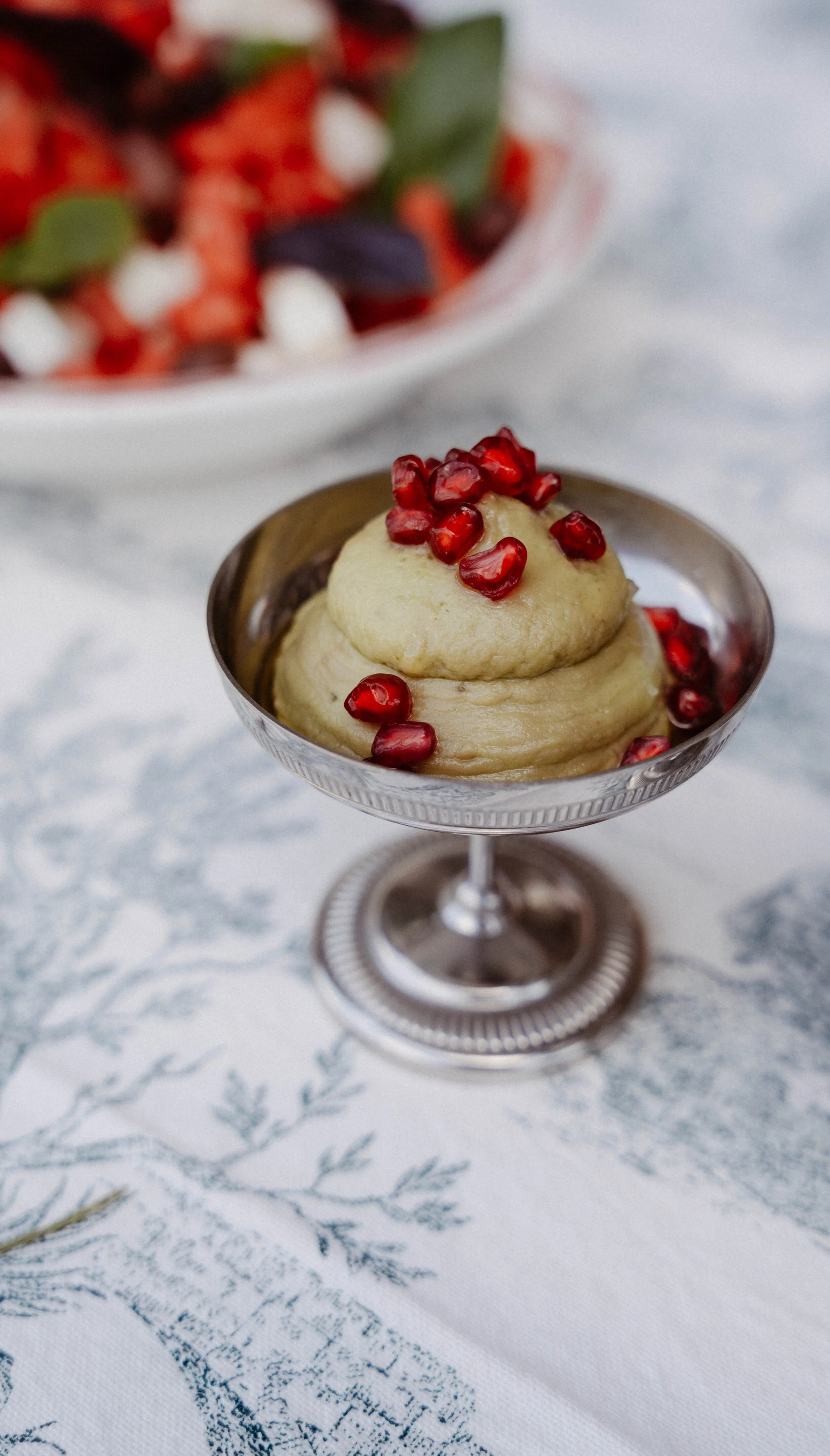 Crème dessert à la vanille garnie de graines de Grenade, servie dans un verre à pied en métal, avec un fond flou de salade de fruits.