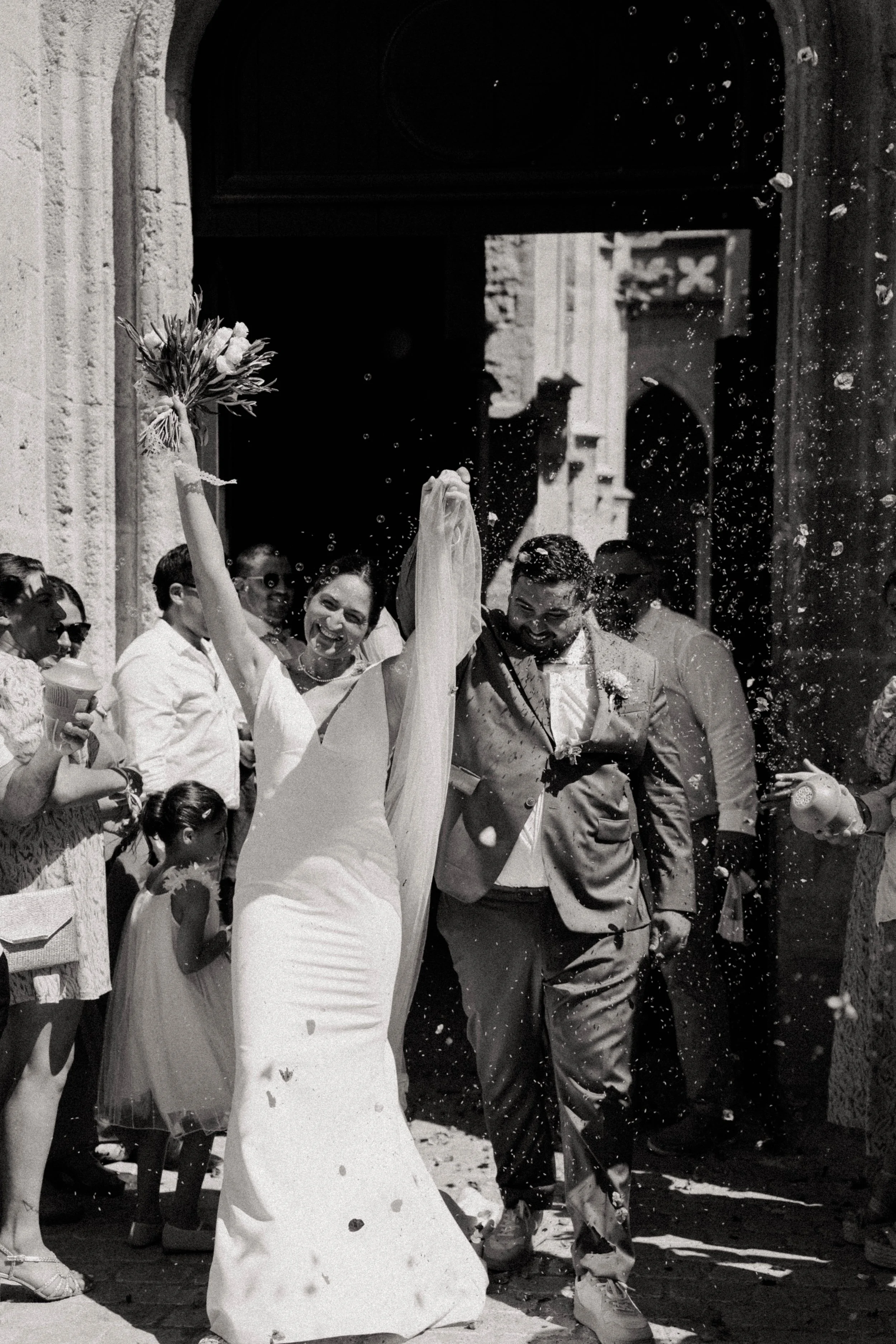 Un mariage avec des invités célébrant à l'extérieur, une femme en robe de mariée souriante, un homme en costume, et des personnes autour, une ambiance festive avec des confettis ou des fleurs.