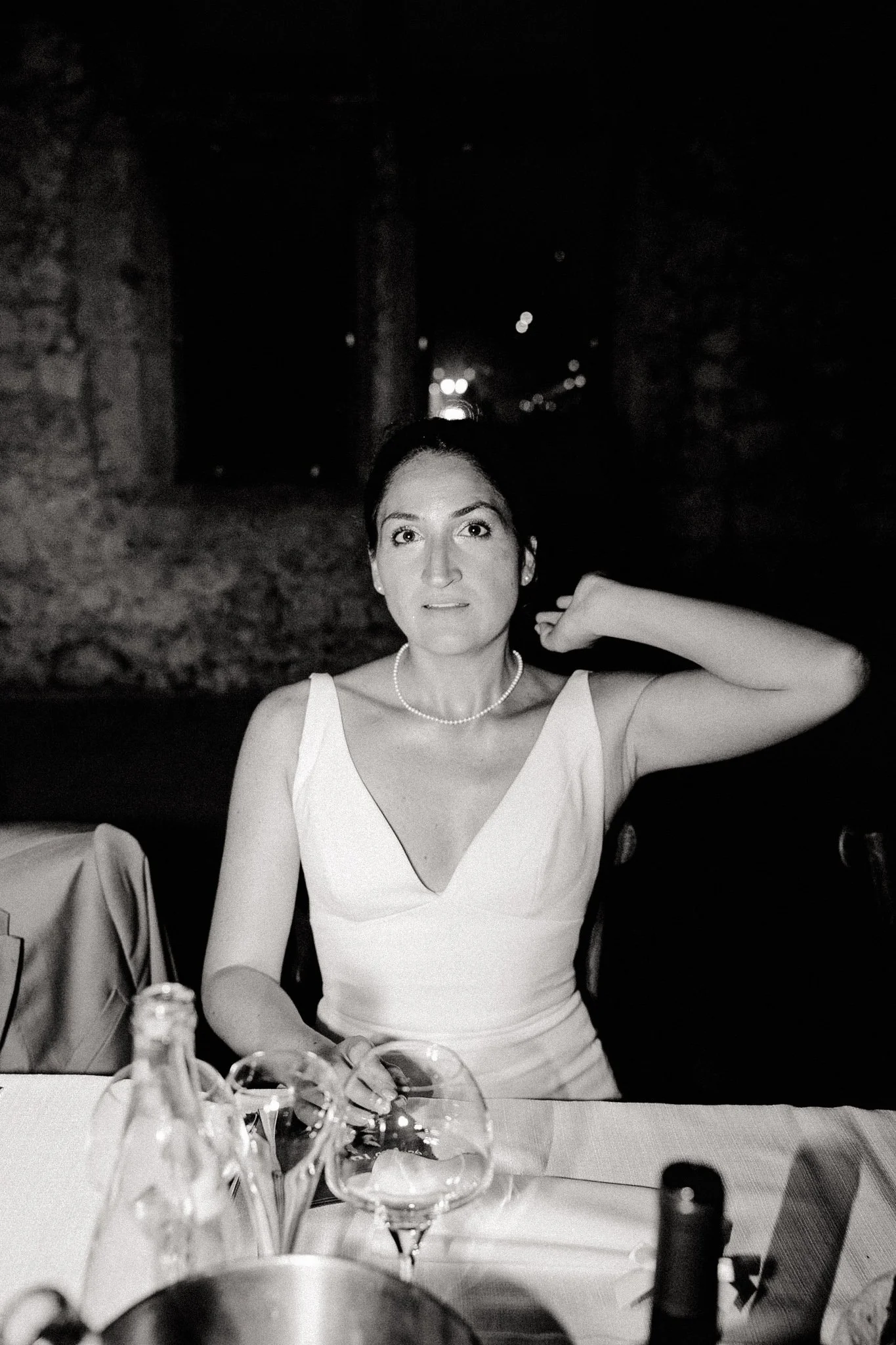 Une femme élégante portant une robe blanche, assise à une table avec des bouteilles et des verres, dans un cadre sombre, probablement lors d'une soirée ou d'un dîner.