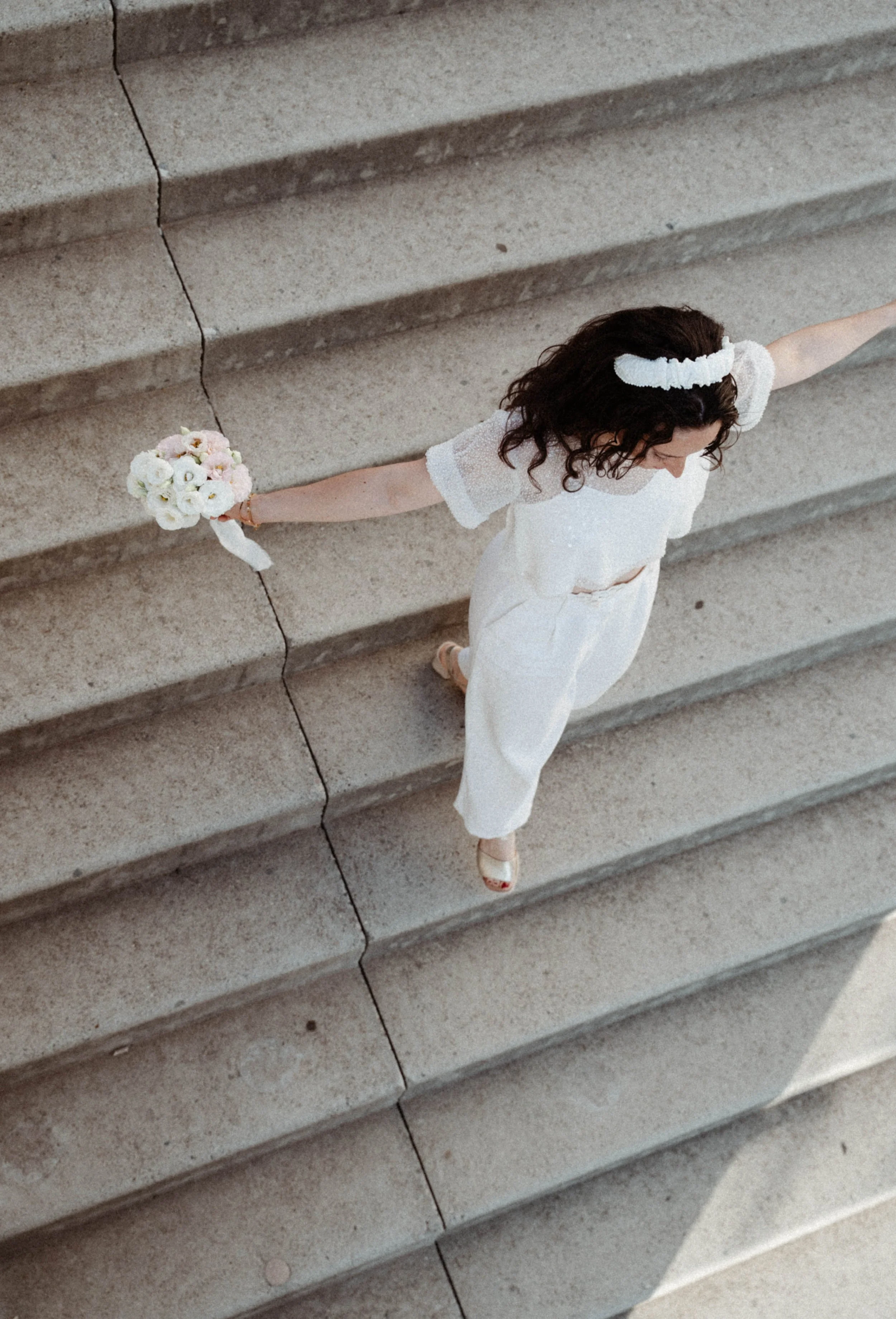 Une femme en robe blanche et avec un bandeau blanc dans les cheveux, tenant un bouquet de fleurs, debout sur des marches en pierre.
