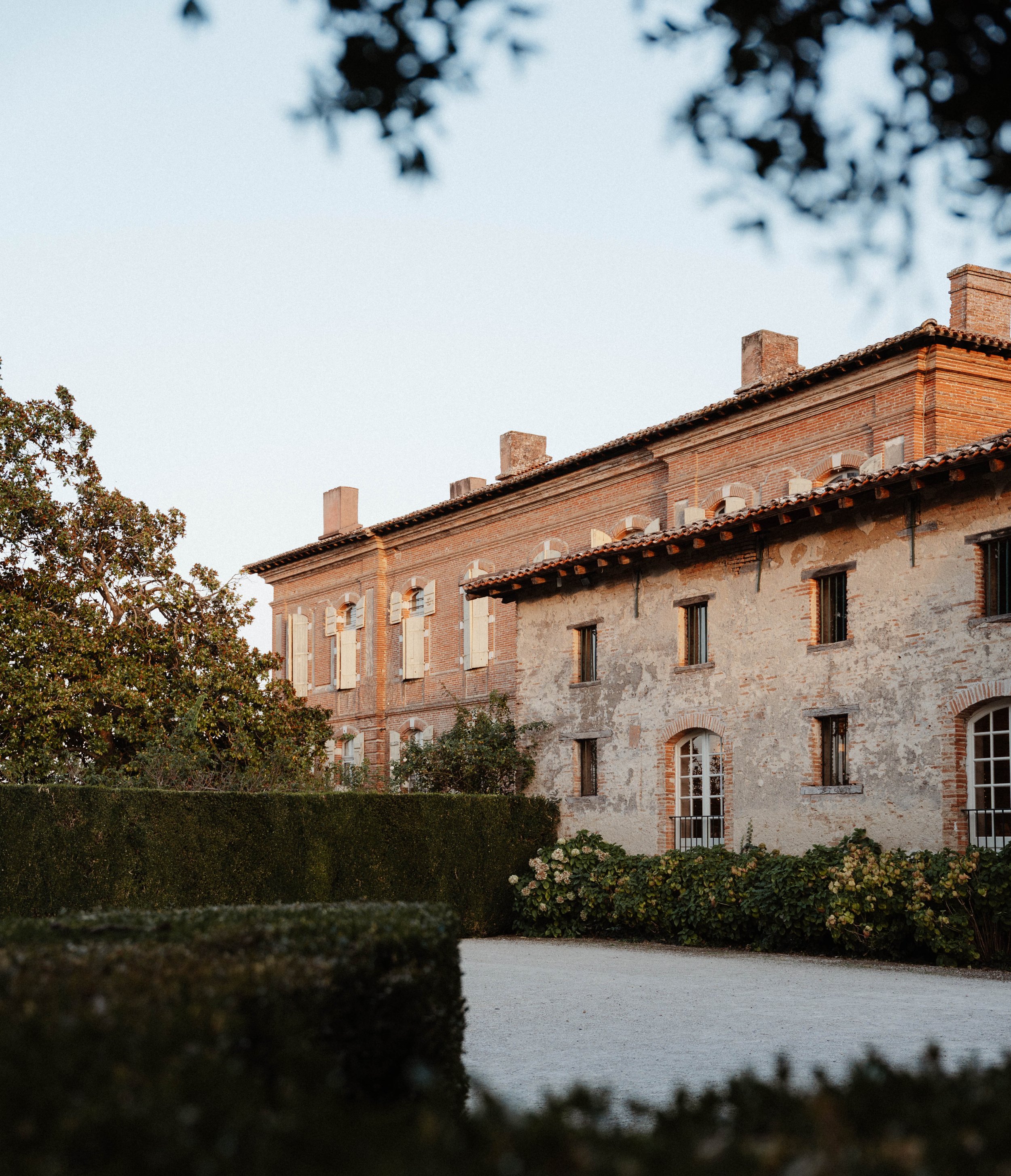 Une vieille maison en pierre avec un toit en tuiles rouges et des fenêtres en arc, entourée d'une haie et d'arbres, avec un ciel clair en arrière-plan.