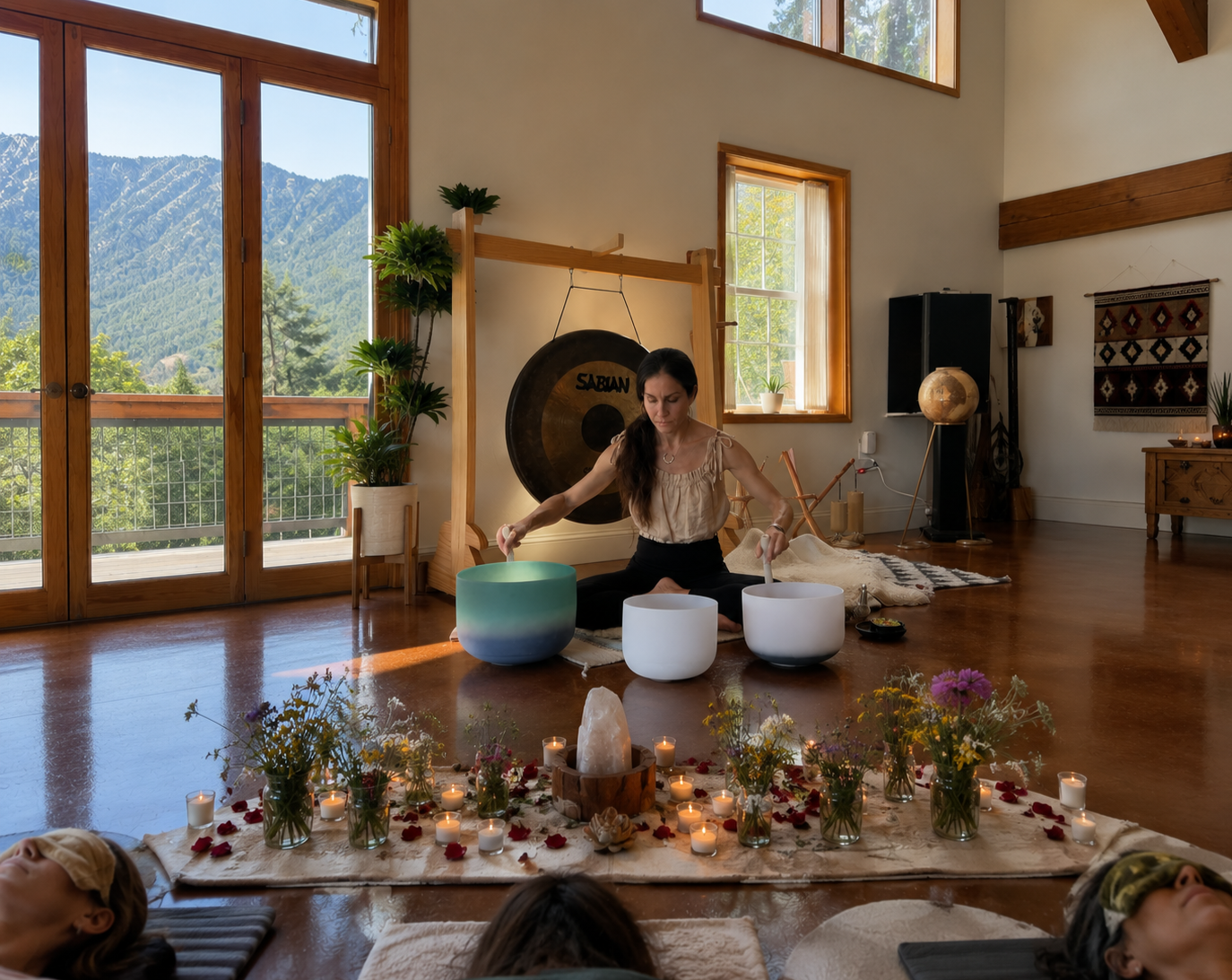 A woman seated on the floor playing crystal singing bowls in a sunlit room with large windows and mountain views. The room is decorated with plants, candles, and a gong, creating a peaceful, meditative atmosphere with a group of people practicing relaxation or meditation.