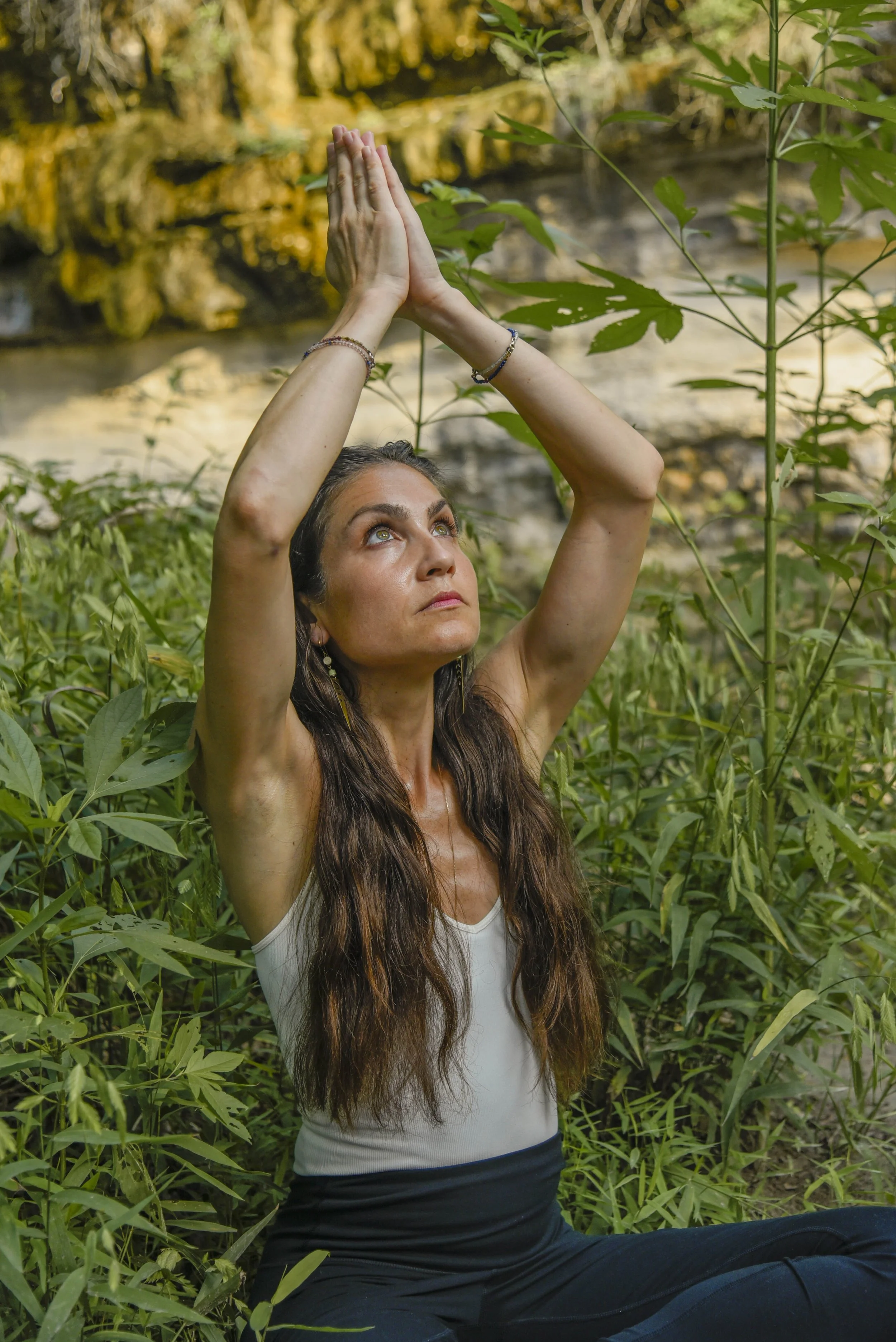 A woman practicing yoga outdoors among tall green plants, with a river and rocks in the background, raising her hands above her head in a yoga pose.