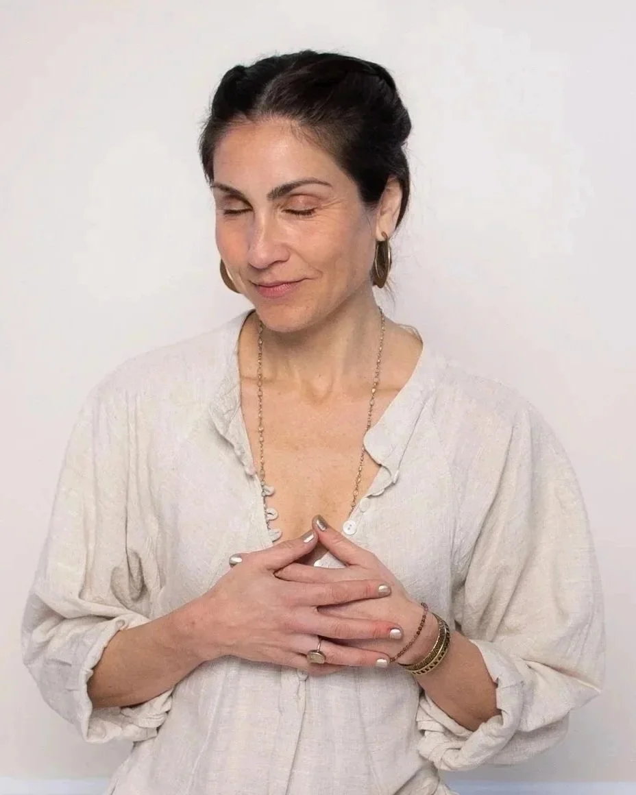 A woman with dark hair pulled back, wearing a beige blouse and jewelry, has her hands placed over her heart and is smiling with her eyes closed.