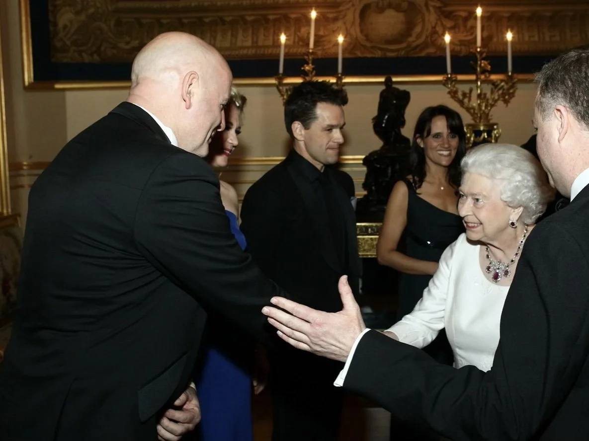Queen Elizabeth II shaking hands with a man at a formal event, with other guests smiling in the background.