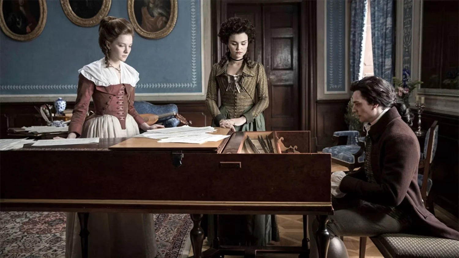 Three people in historical clothing gathered around a piano in an ornate room, with two women standing and a man sitting at the piano.