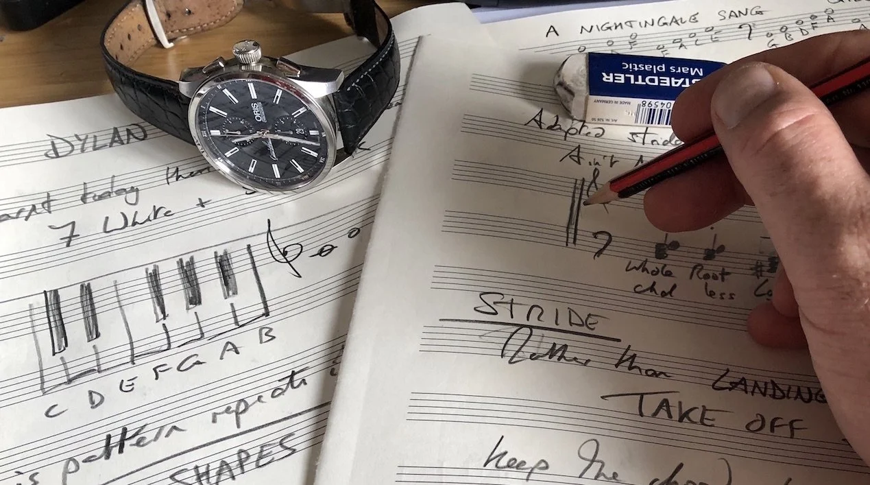 A person's hand writing musical notes on sheet music with a red pencil, with a wristwatch placed on another sheet, and an eraser, cap, and sheet music scattered on a wooden surface.