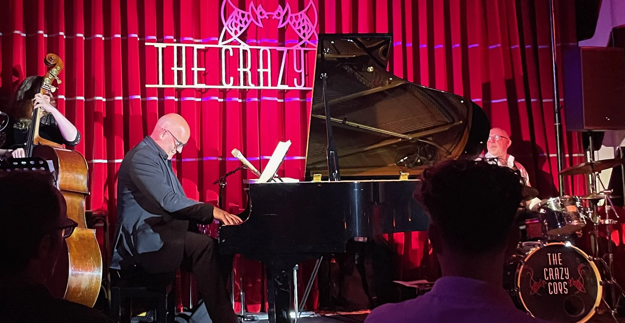 Jazz band performing on stage with a red curtain backdrop, including a pianist, a bassist, and a drummer, at The Crazy Coqs venue.