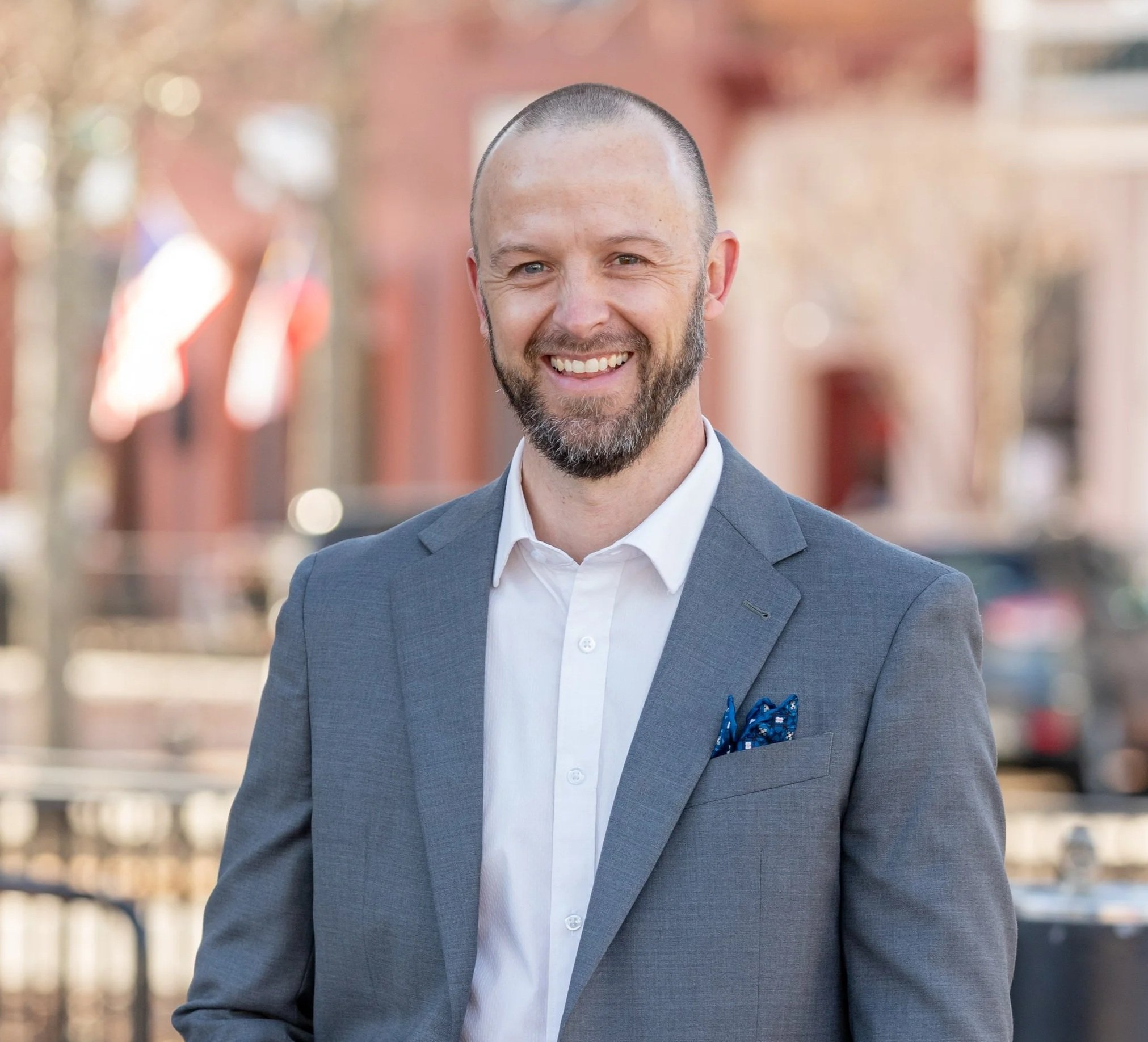 A smiling man with a beard and a bald head, dressed in a gray suit and white shirt, outdoors in an urban area with blurred background of buildings and colorful decorations.