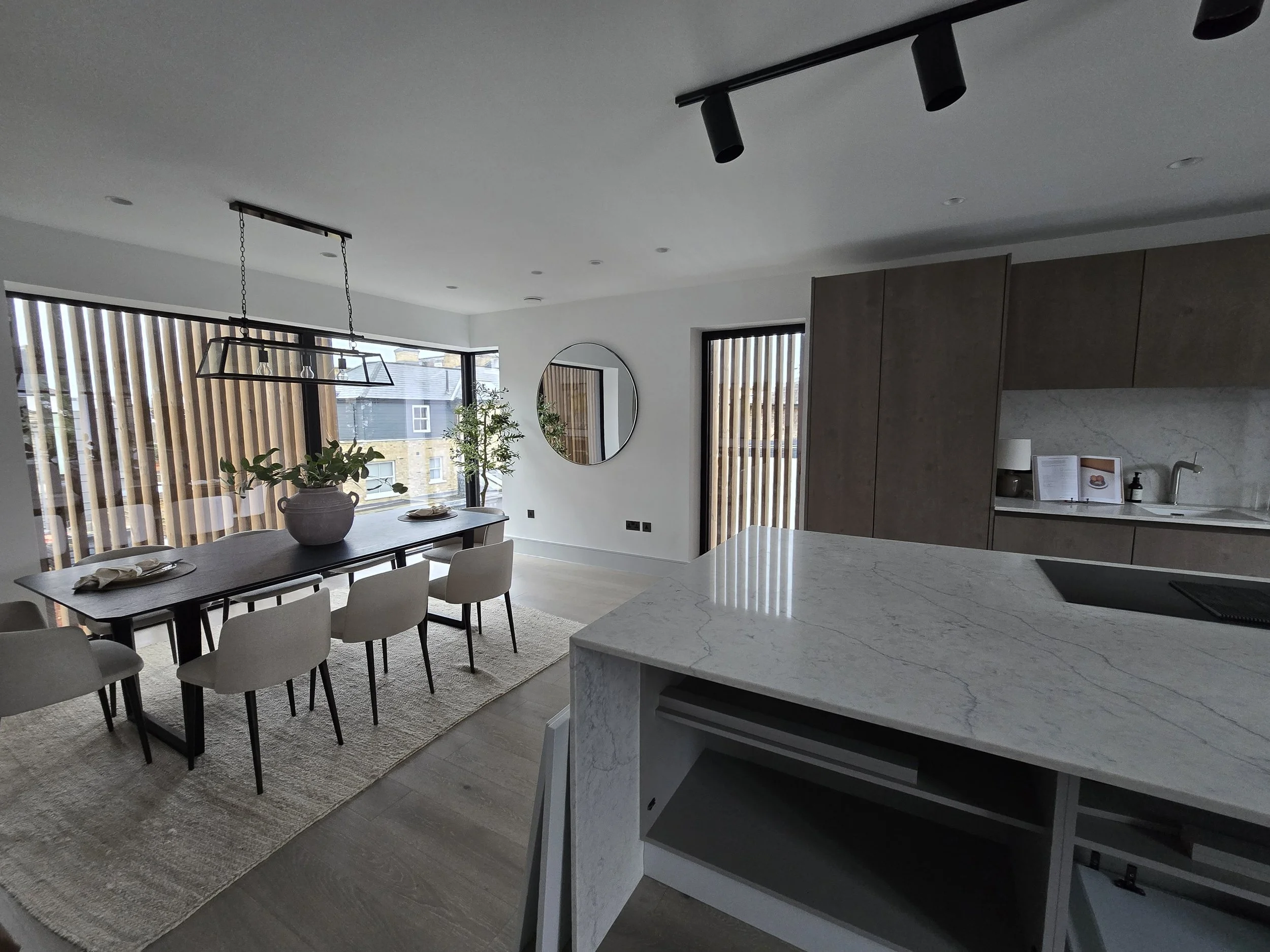 Modern kitchen and dining area with large windows, wooden blinds, and minimalist decor, including a marble kitchen island.