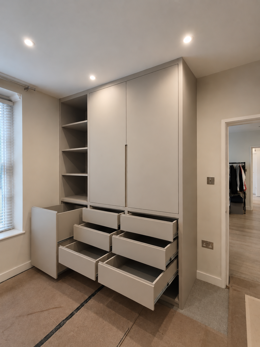 A modern built-in gray wardrobe with open drawers and shelves in a room with beige walls, ceiling lights, and a window with blinds.