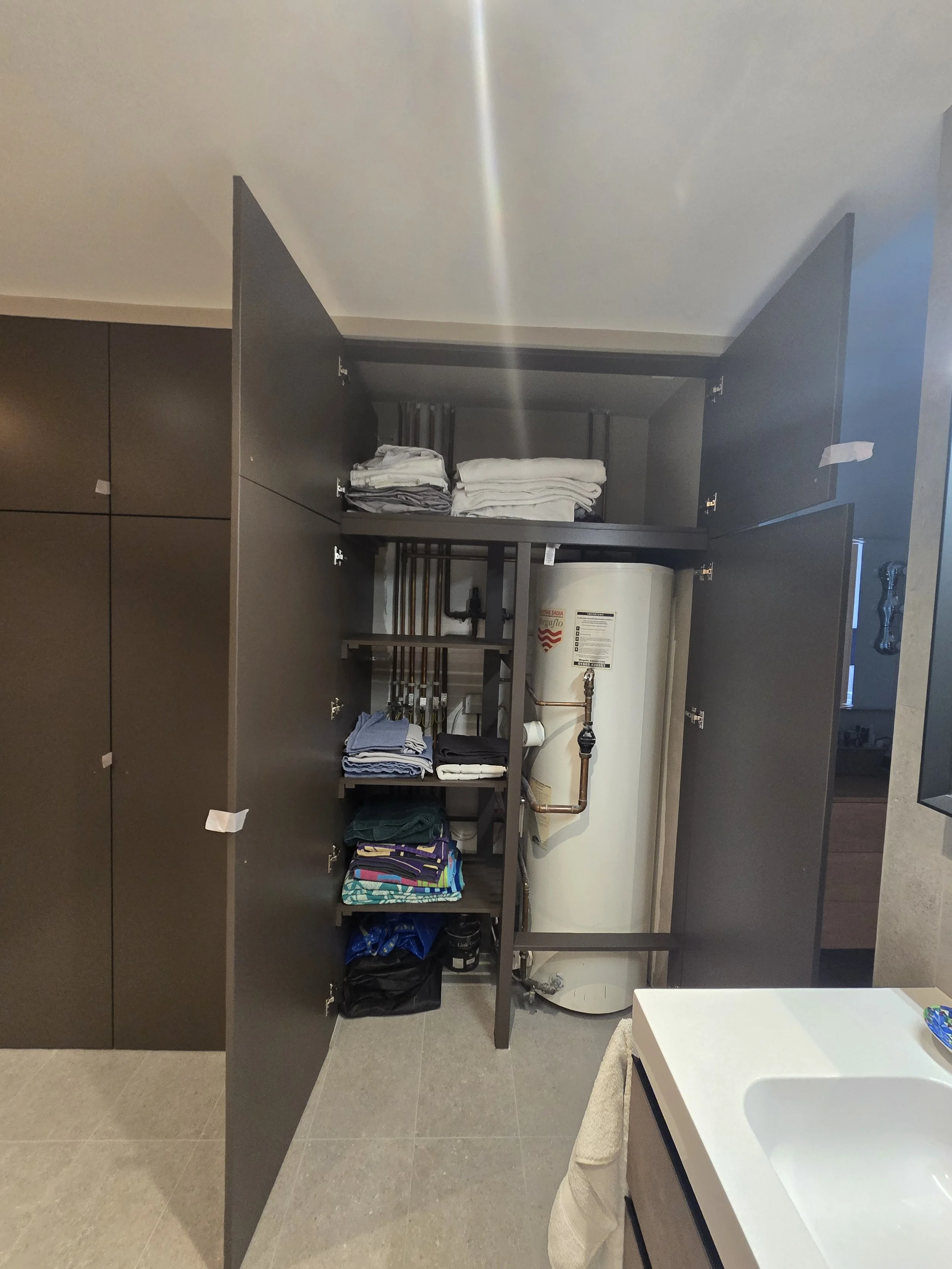 Open bathroom cabinet with folded towels, laundry, and a water heater inside, adjacent to a bathroom sink with a towel underneath.