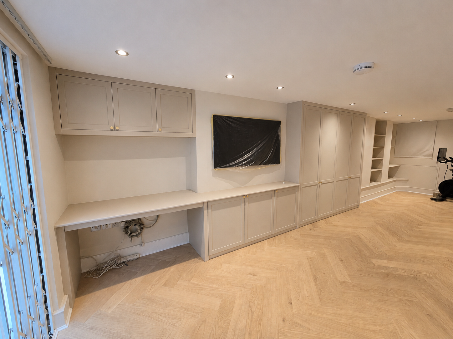 Empty room with built-in beige cabinets, a wall-mounted television covered with plastic, and an exercise bike in the corner. Light hardwood floors and recessed ceiling lights.