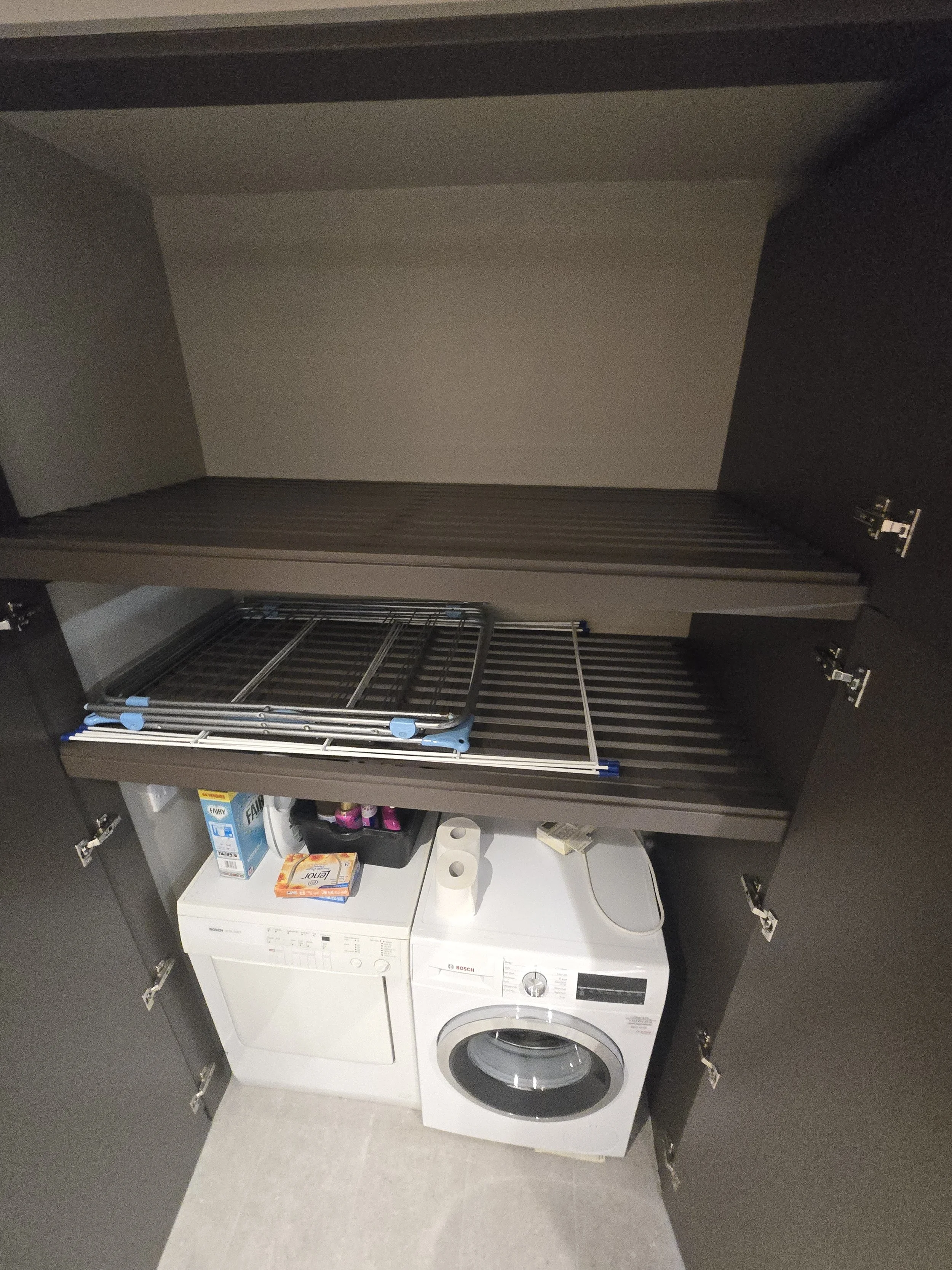 A laundry room with a white washing machine, black cupboards, and a foldable drying rack. Items like a box of laundry detergent, a box of FabriClear, and some pink spray bottles are on top of the washing machine.