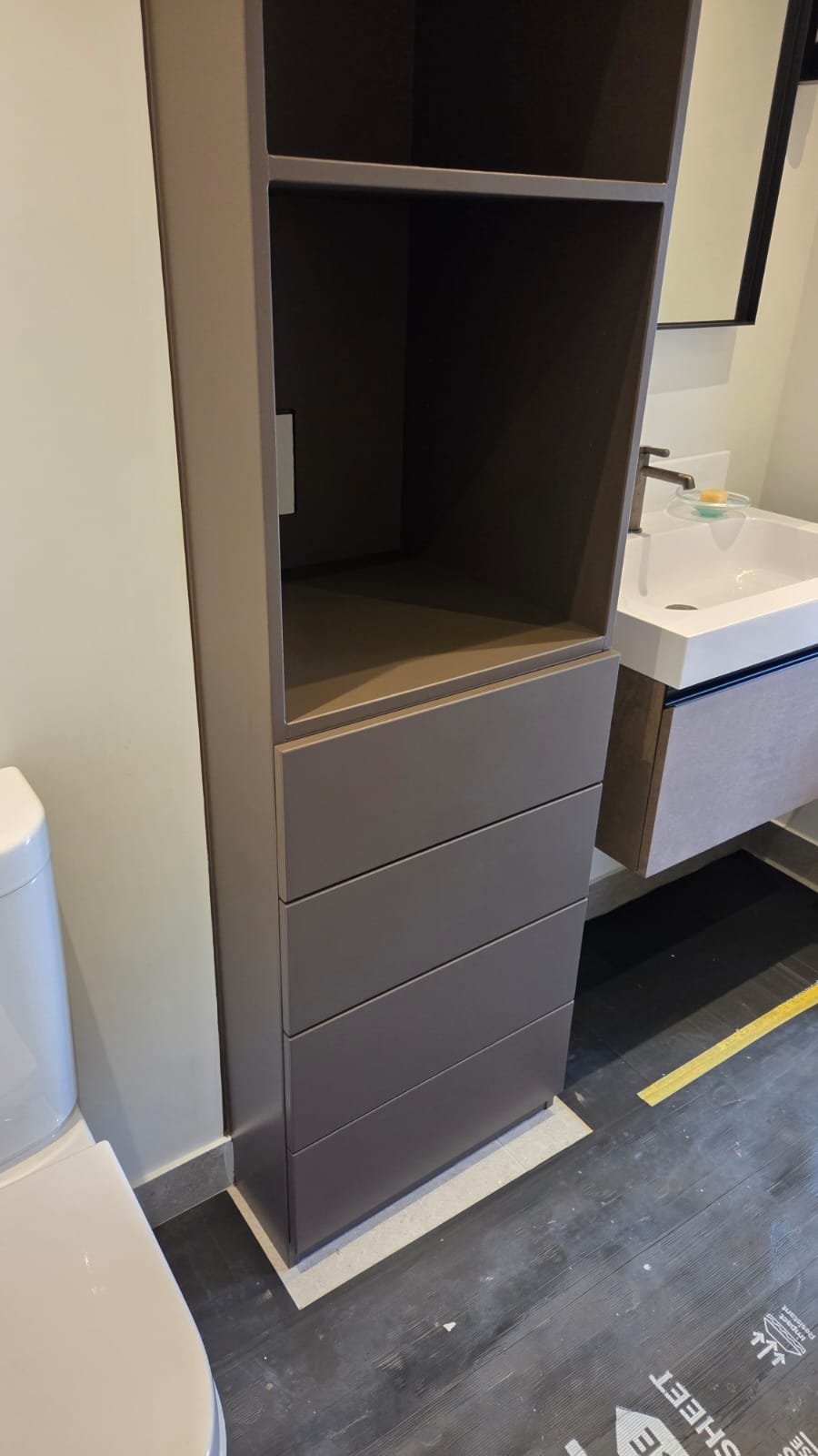 Brown cabinet with open upper shelf and four drawers below, located next to a white bathroom sink and partial view of a white toilet.