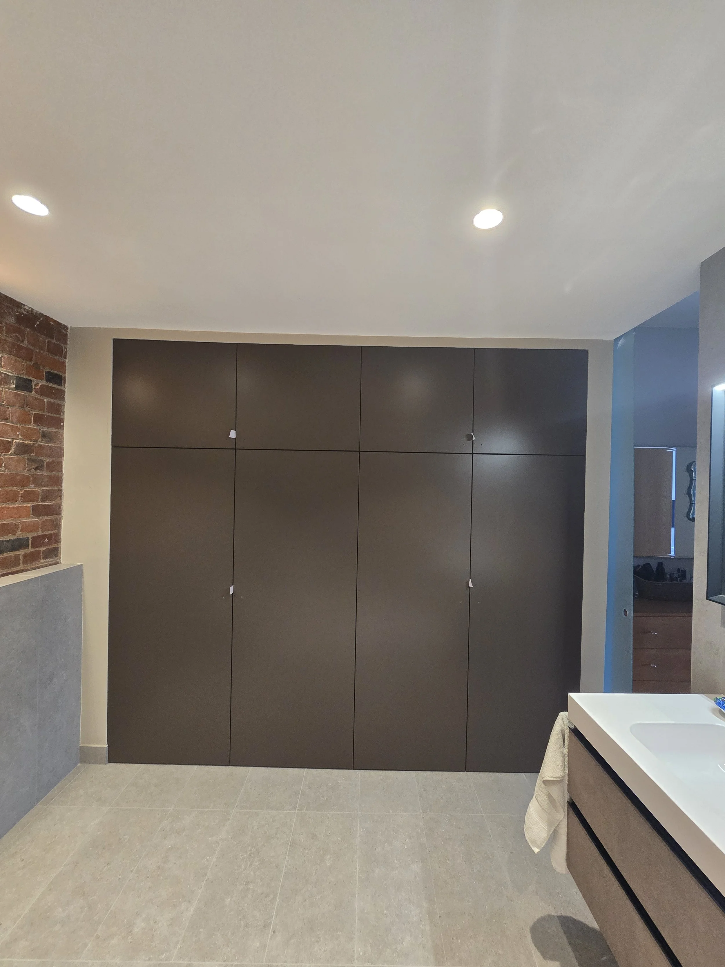 Modern bathroom with gray tiled floor, black cabinets, a white countertop with a sink, a towel hanging below, a blue wall on the right, a brick wall on the left, and recessed ceiling lights.
