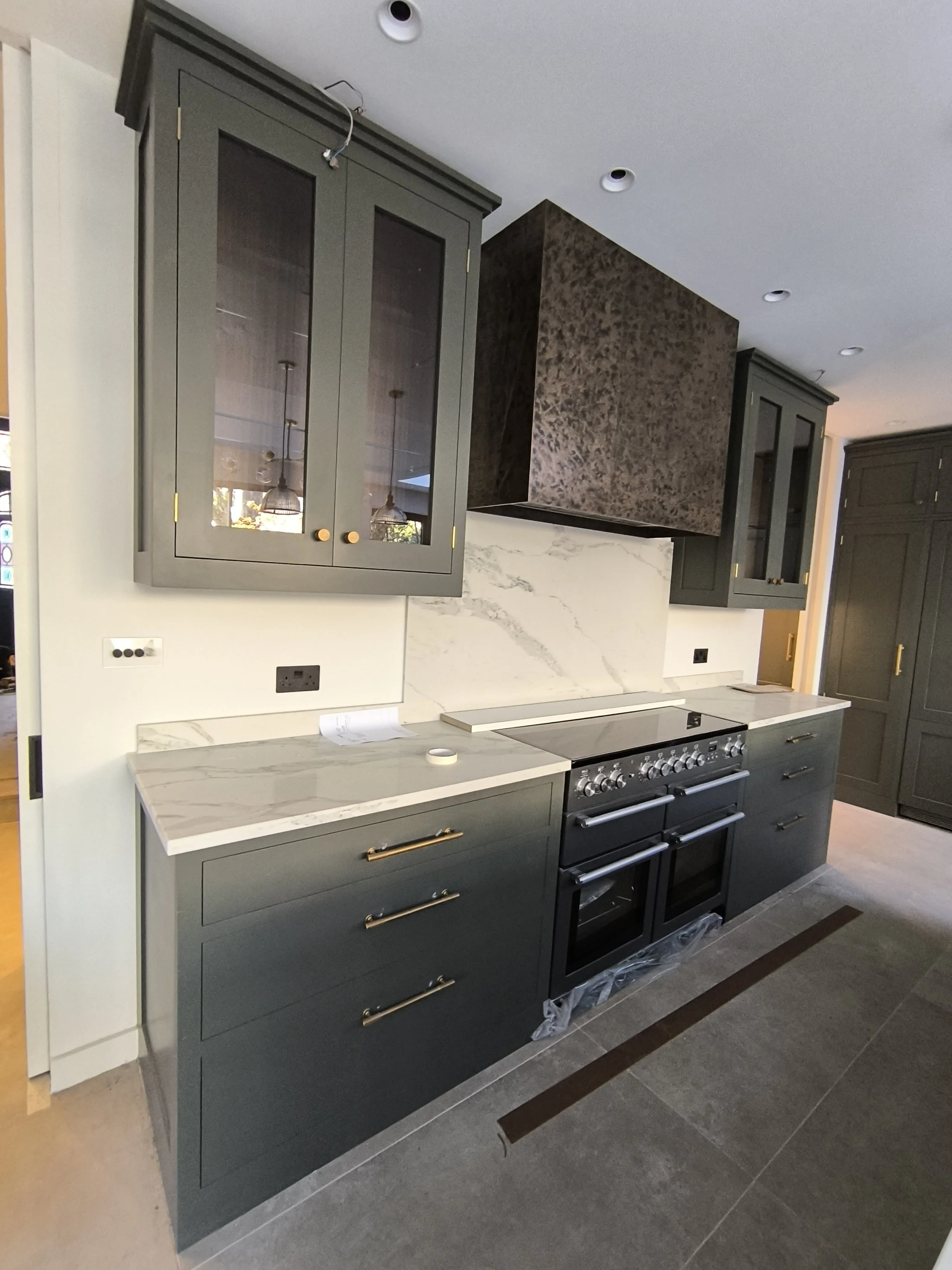 Modern kitchen with dark gray cabinets, a marble countertop, and a matching marble backsplash. There is a built-in oven and stove, with some protective plastic wrap still on parts of the appliances. Overhead are recessed ceiling lights, and a partial