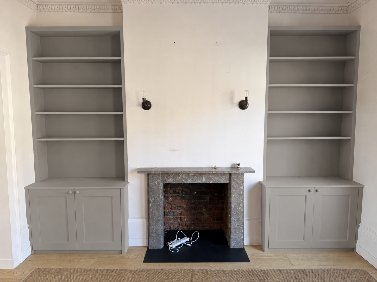 Empty fireplace with a marble mantel and two empty built-in bookshelves on either side, wall-mounted light fixtures above the fireplace, and electrical cords on the floor in front.