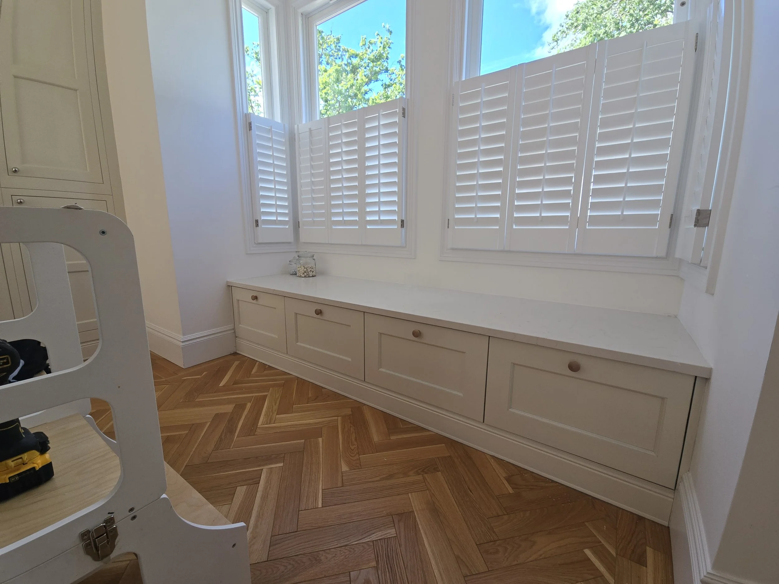A bright room with white walls and a large window featuring white plantation shutters. Outside the window, green trees and a blue sky are visible. The room has a built-in white cabinet with drawers and a marble countertop. Part of a white wooden chai