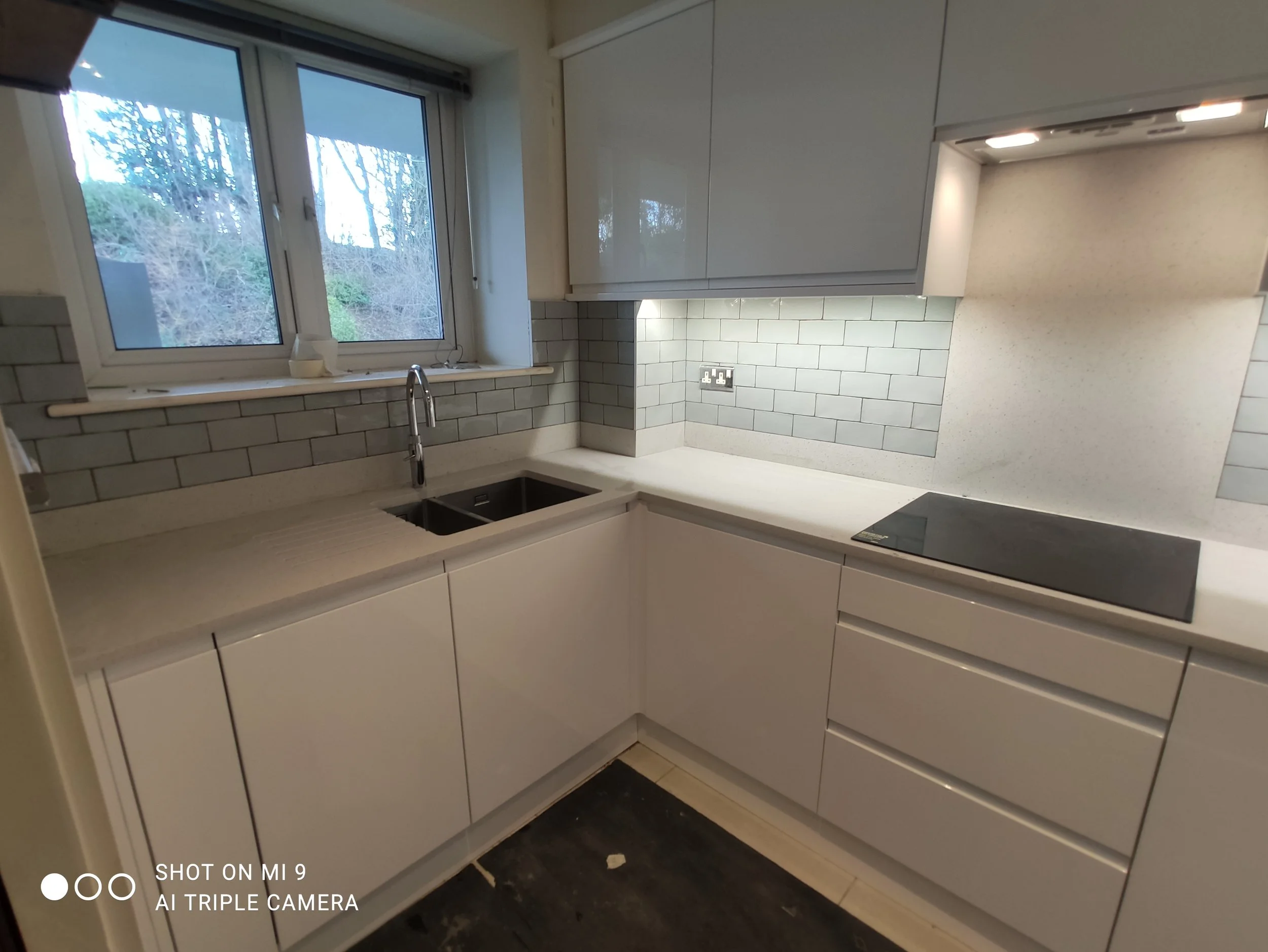 A kitchen with white cabinets, a window showing trees outside, a gray subway tile backsplash, and a black tile floor. There is an oven underneath the counter on the right side.