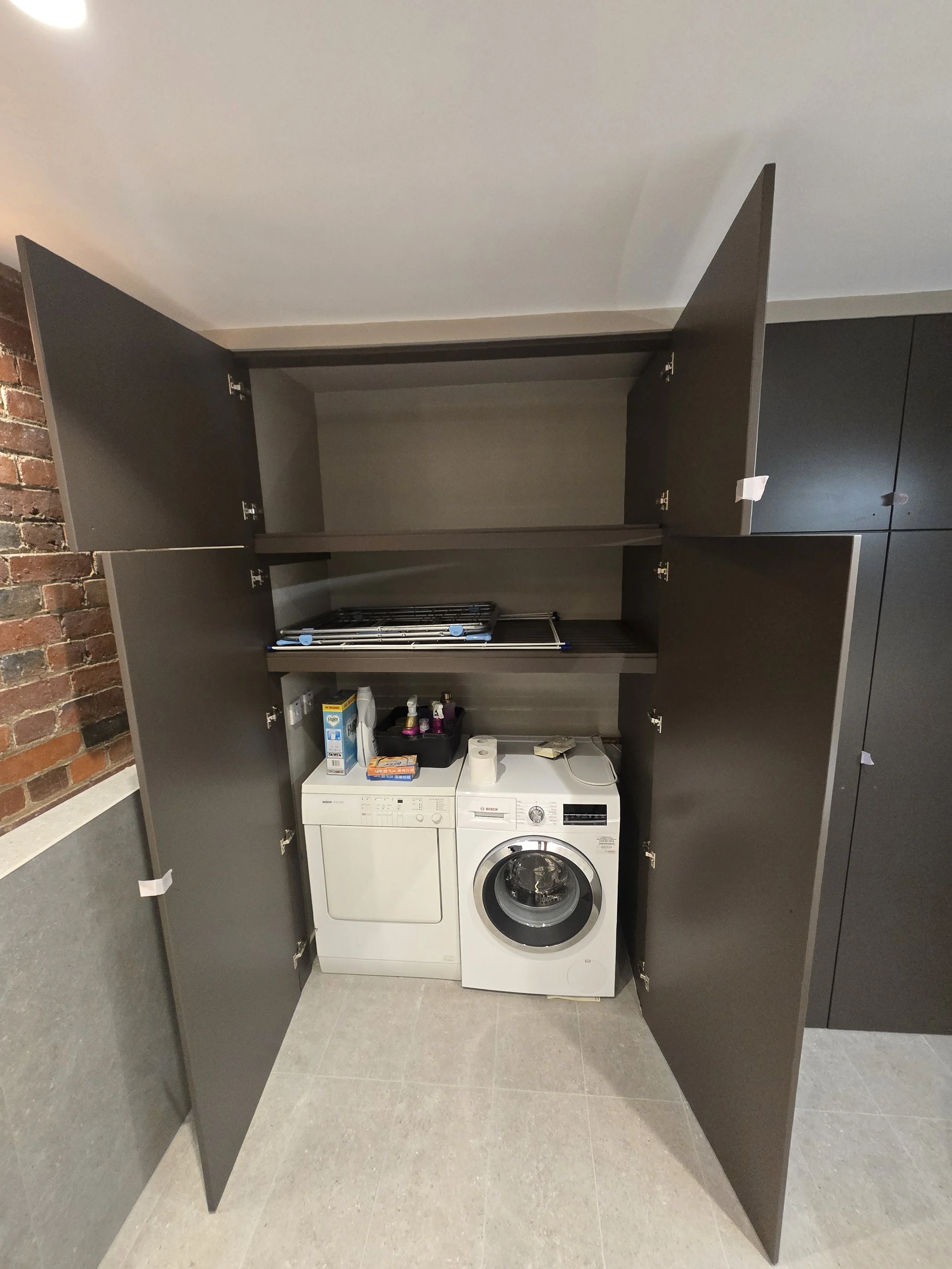A washer and dryer set inside an open dark-colored cabinet with storage space above, with laundry supplies on top, in front of a brick wall and light-colored flooring.