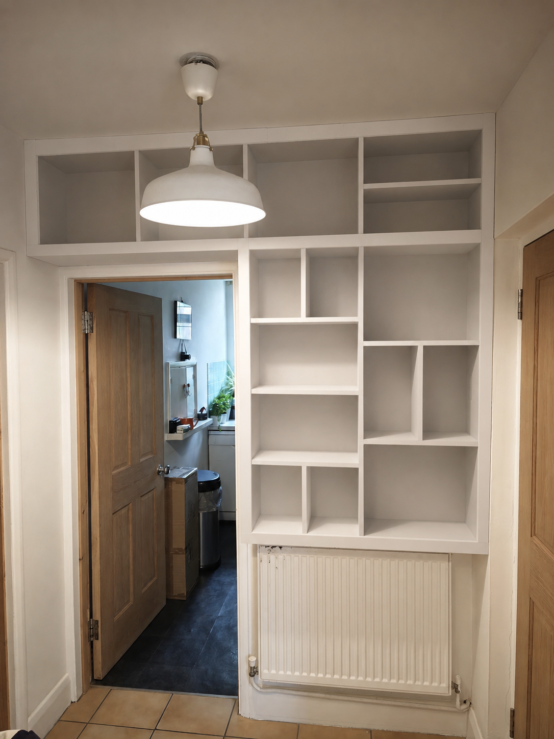 Empty white built-in shelving unit and radiator in a doorway leading to a room with a washing machine and window with plants.