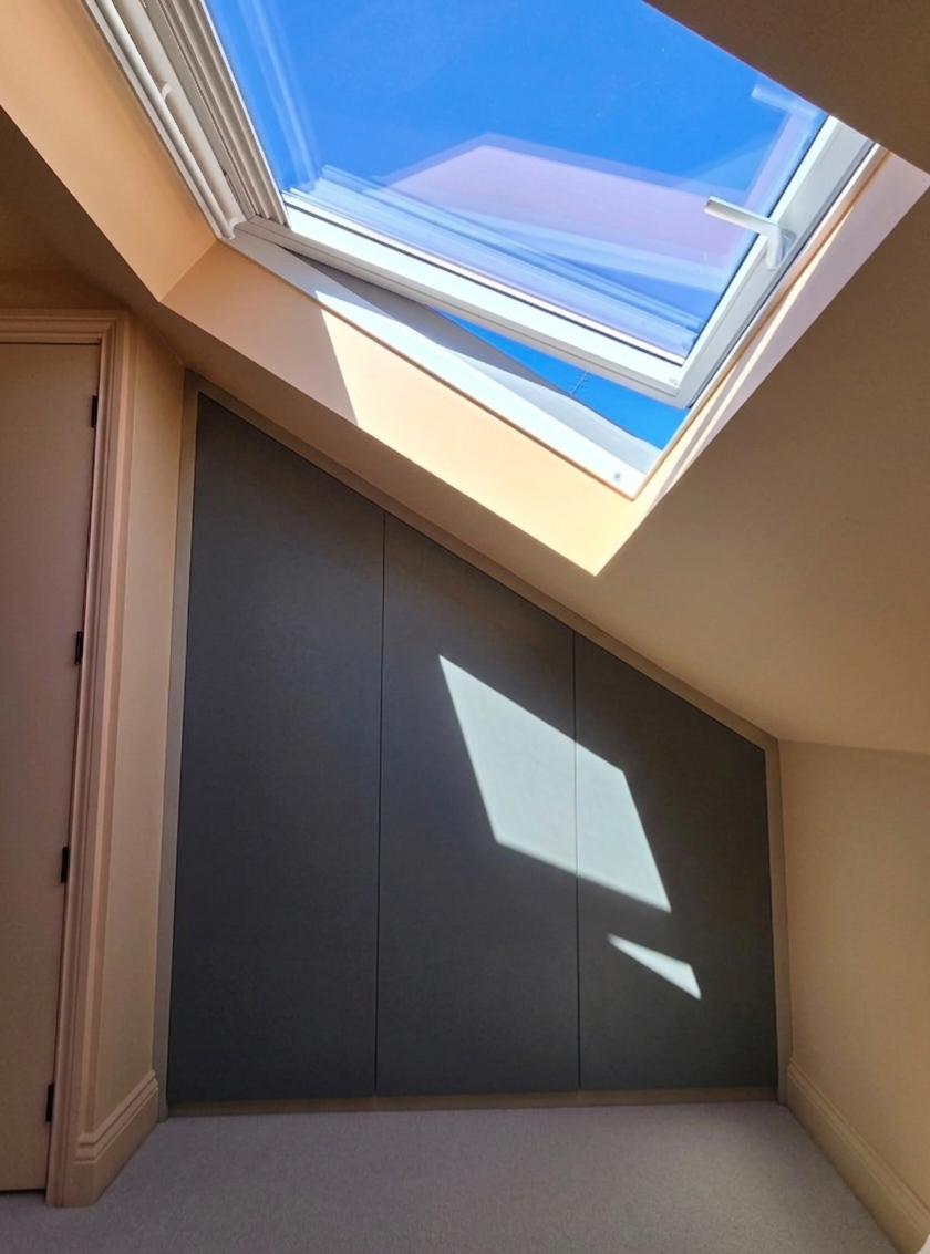 A room with a sloped ceiling featuring an open skylight window. The sky outside is clear and blue, and sunlight casts a shadow of the window onto the wall below.