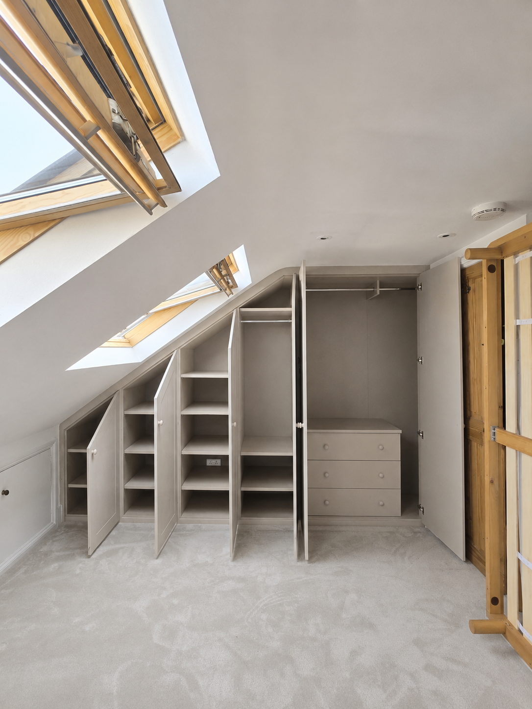 Empty attic room with sloped ceiling and skylight windows, built-in grey wardrobe with open doors and drawers, and light-colored carpet flooring.