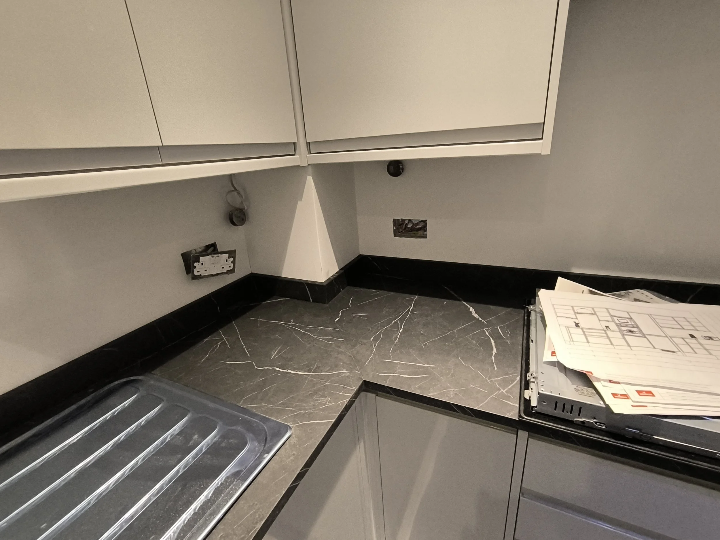 Unfinished kitchen with gray cabinets, black marble countertop, and a laundry sink, with electrical outlets and a blueprint on the counter.