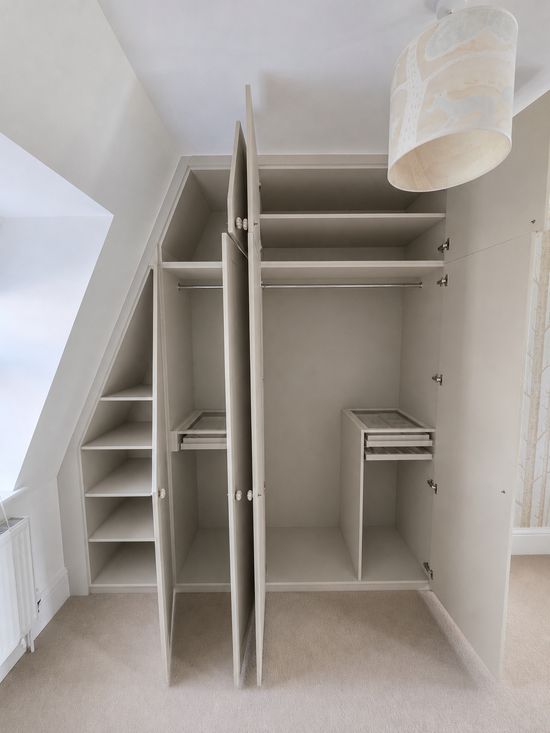 Open built-in white wardrobe with shelves and hanging space in an attic room. A window with sloped ceiling is visible, with beige carpeted floor and patterned wallpaper on adjacent wall.