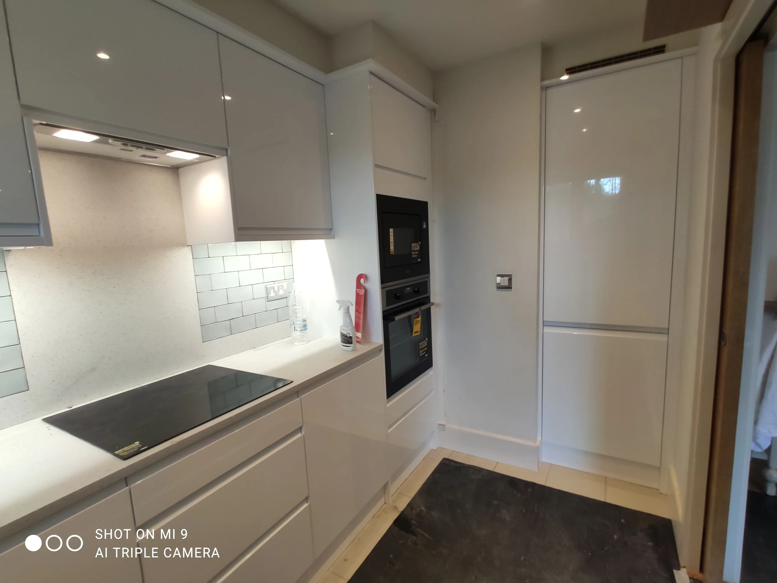 Modern kitchen with white cabinets, a black induction cooktop, and built-in oven. White subway tile backsplash and a countertop with cleaning spray and a water bottle.