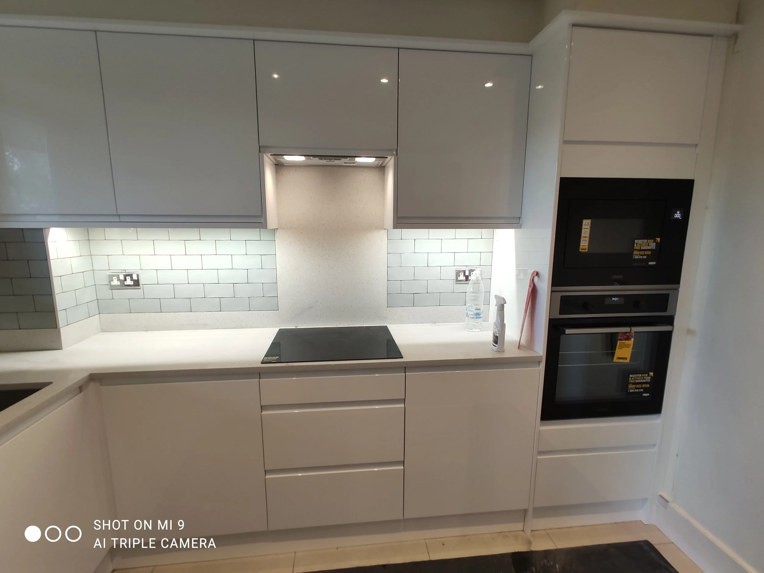A modern kitchen with white cabinets, a built-in oven, a microwave, a black electric cooktop, and a backsplash with white subway tiles. There is a clear water bottle and a spray bottle on the counter.