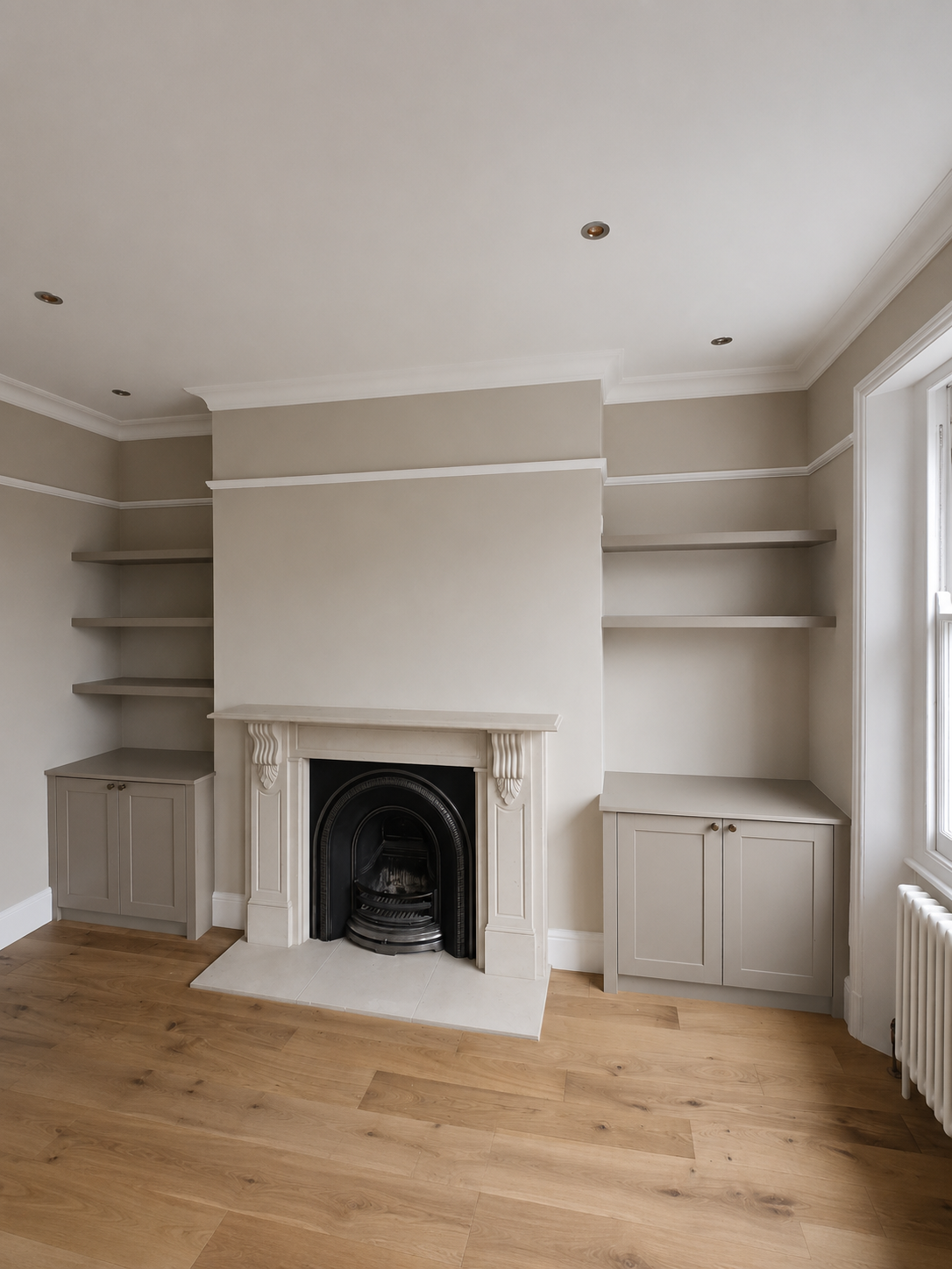 Empty living room with beige walls, white crown molding, built-in bookshelves on either side of a fireplace, and a window on the right. Hardwood floors and a white radiator below the window.